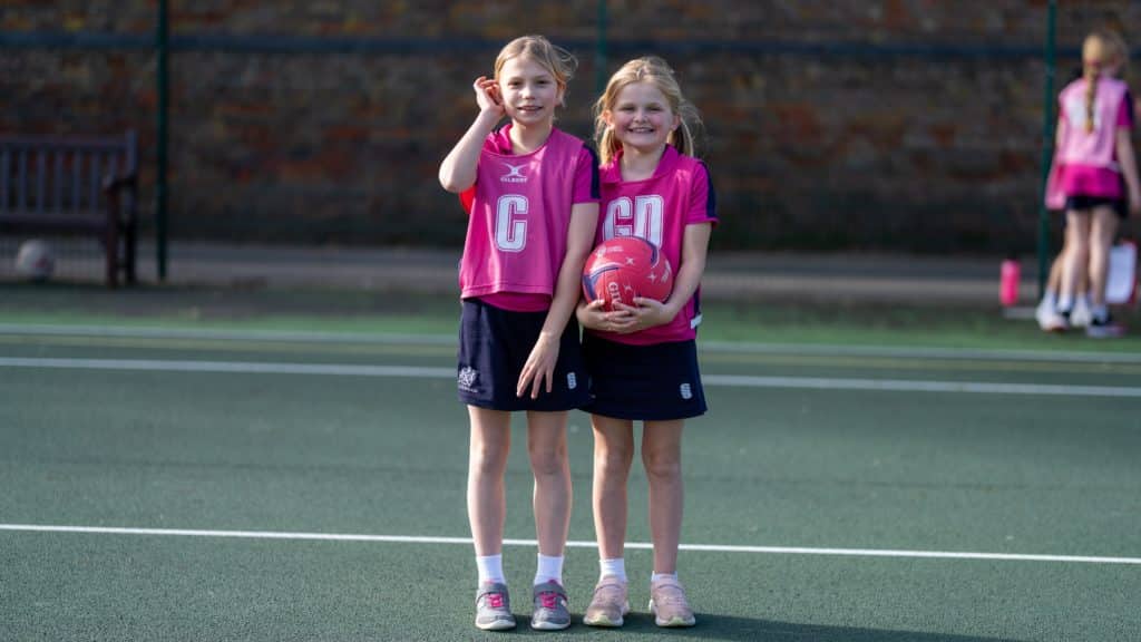 Two young girls in sports kit stand on an outdoor court at Cheltenham Prep School. One girl is holding a pink netball, and both are smiling at the camera. Two other children are visible in the background, near a bench.