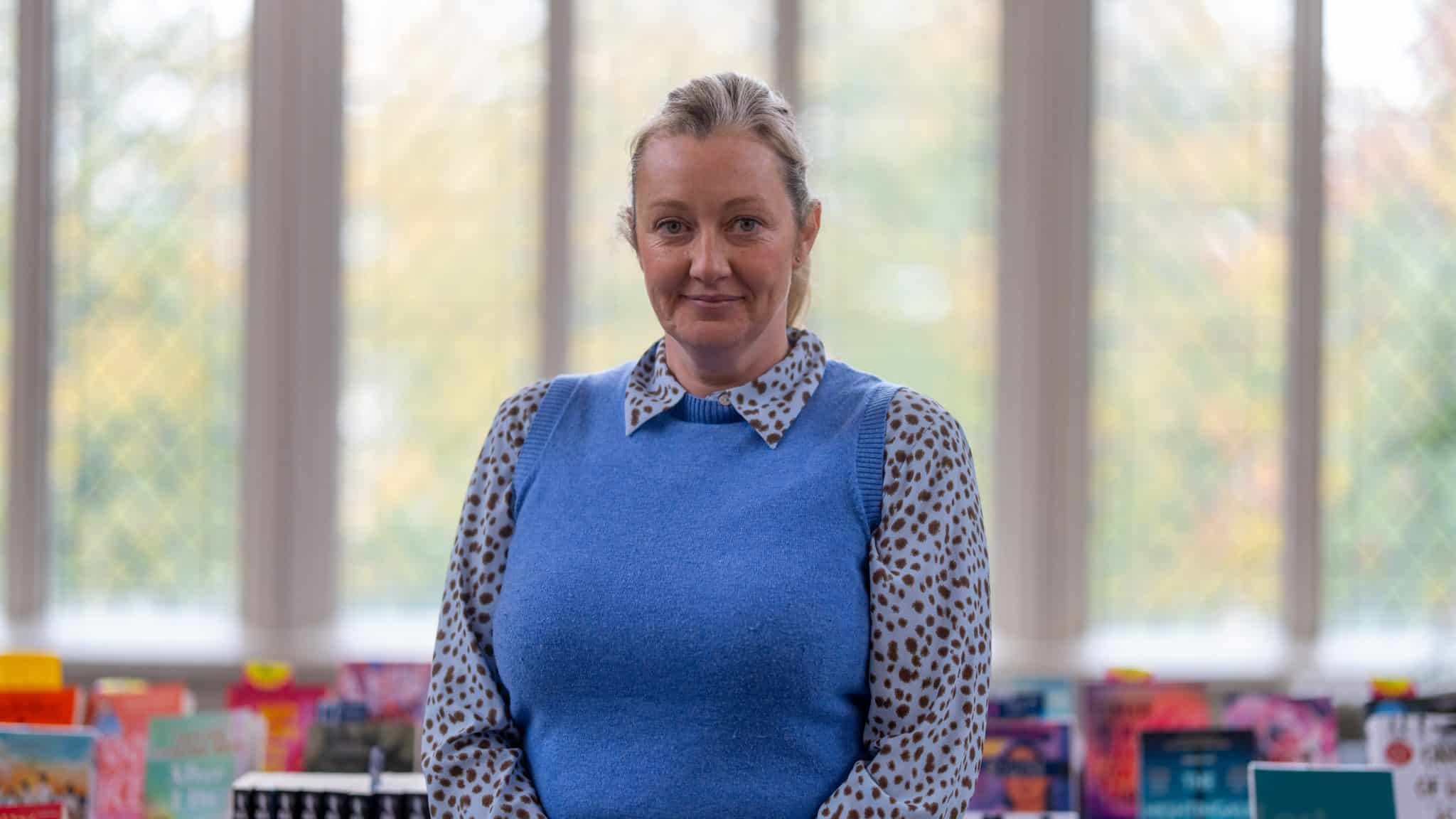 A woman with blonde hair tied back, wearing a blue waistcoat over a spotted shirt, stands indoors in front of large windows, perhaps awaiting Cheltenham College admissions, with a blurred background of books and greenery outside.