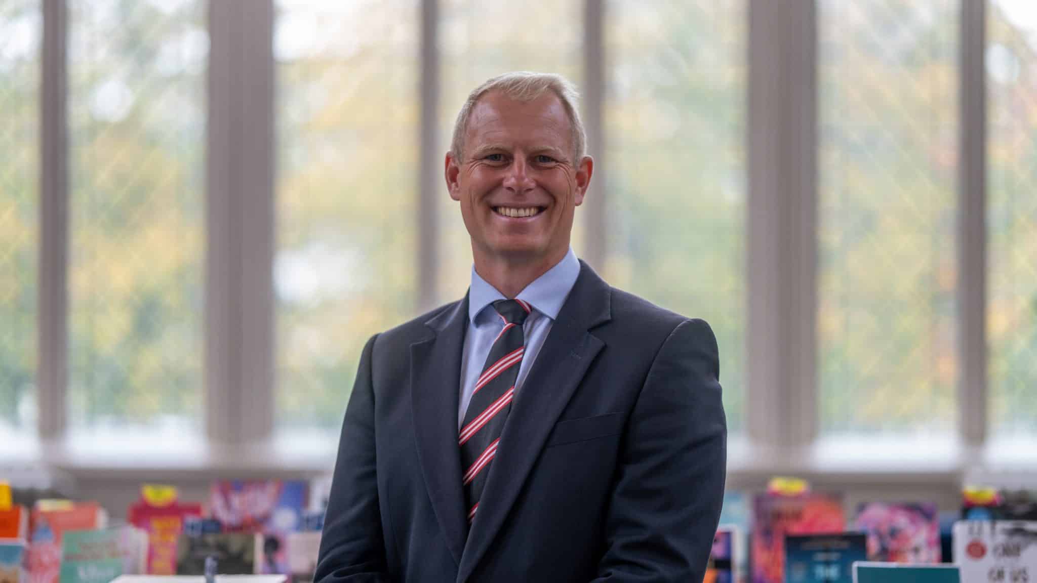 A man in a suit and striped tie is smiling, standing indoors in front of large windows with diamond-shaped panes—an inviting scene that reflects the welcoming spirit of Cheltenham College admissions. Shelves with colourful books are visible in the blurred background.