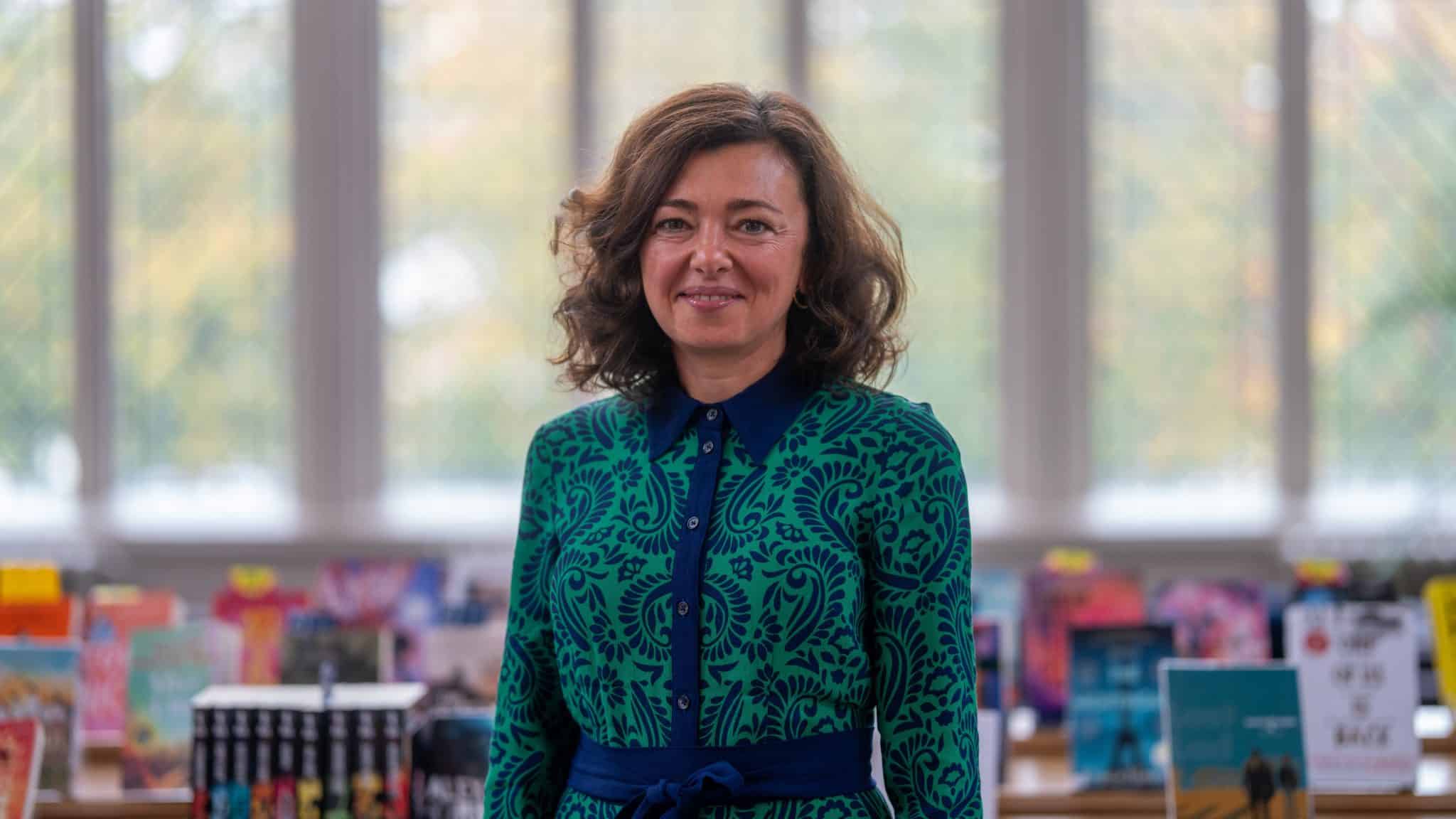 A woman with wavy brown hair wearing a green and blue patterned dress stands smiling in front of a blurred background of books, large windows, and Cheltenham College admissions materials on display.