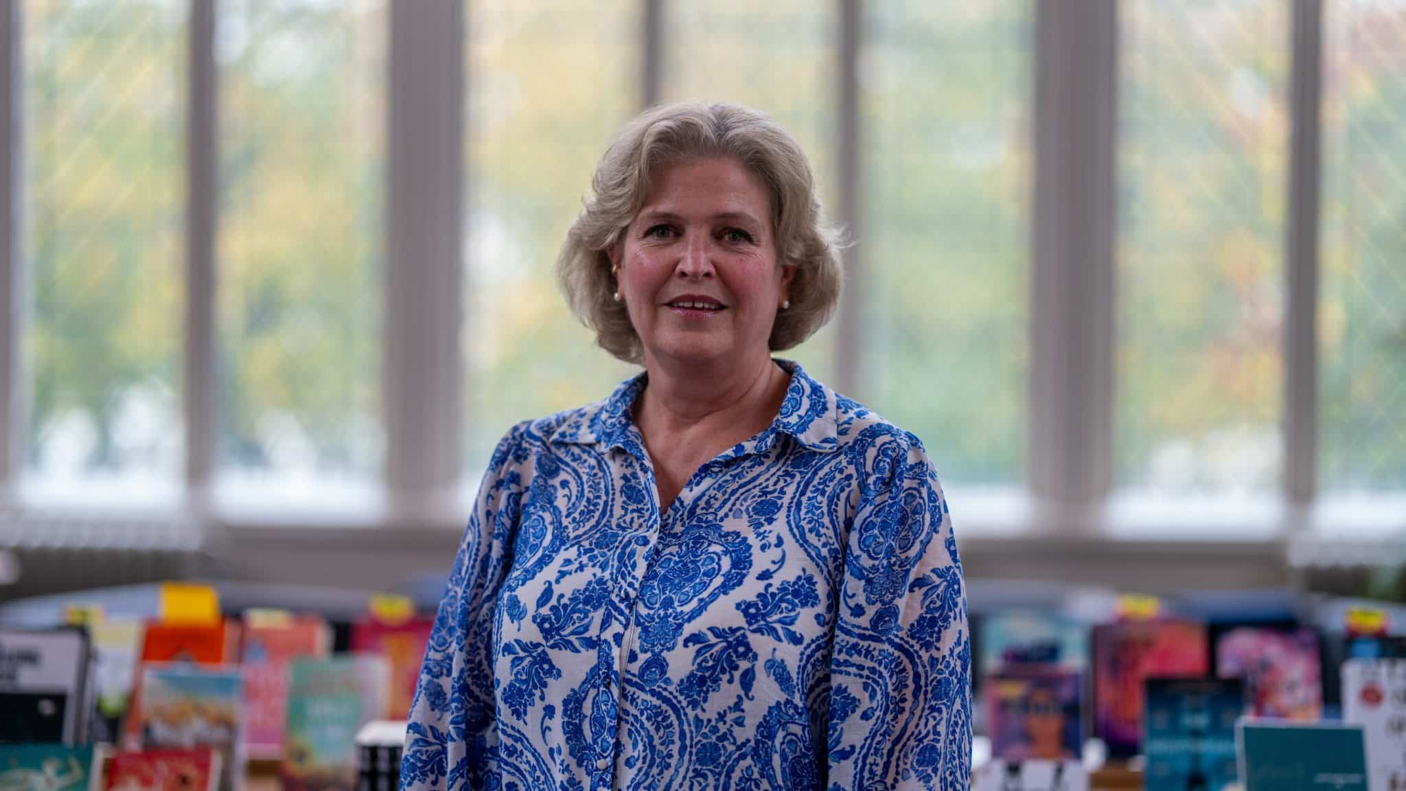 A woman with short, curly blonde hair wearing a blue and white patterned blouse stands in front of a large window with books displayed on tables behind her, evoking an inviting atmosphere reminiscent of Cheltenham College admissions. The background is softly blurred.