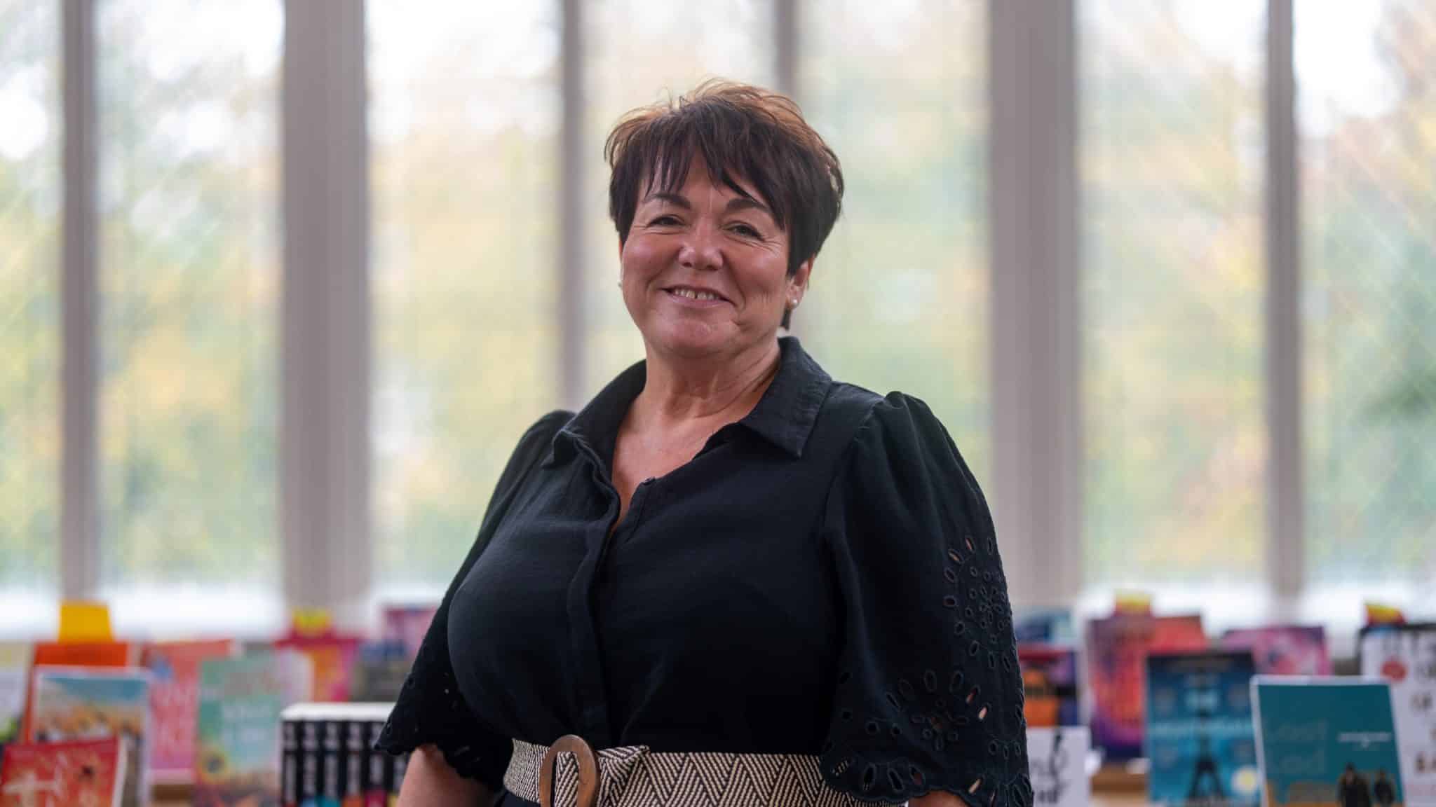 A woman with short dark hair and a black blouse stands smiling in front of shelves filled with colourful books, as natural light pours through large windows—reflecting the welcoming atmosphere of Cheltenham College admissions.