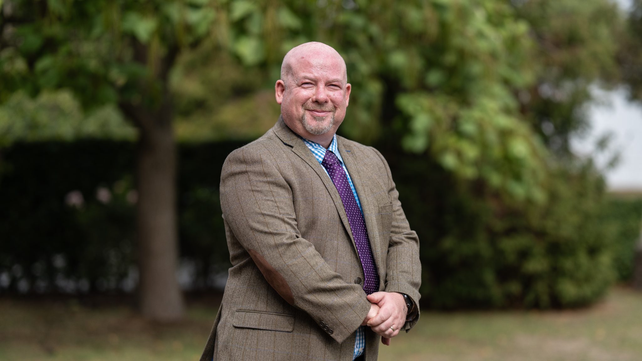 A bald man with a goatee, wearing a brown suit, blue chequered shirt, and purple tie, stands outdoors smiling—an approachable face for Cheltenham College admissions—with green trees and blurred foliage in the background.