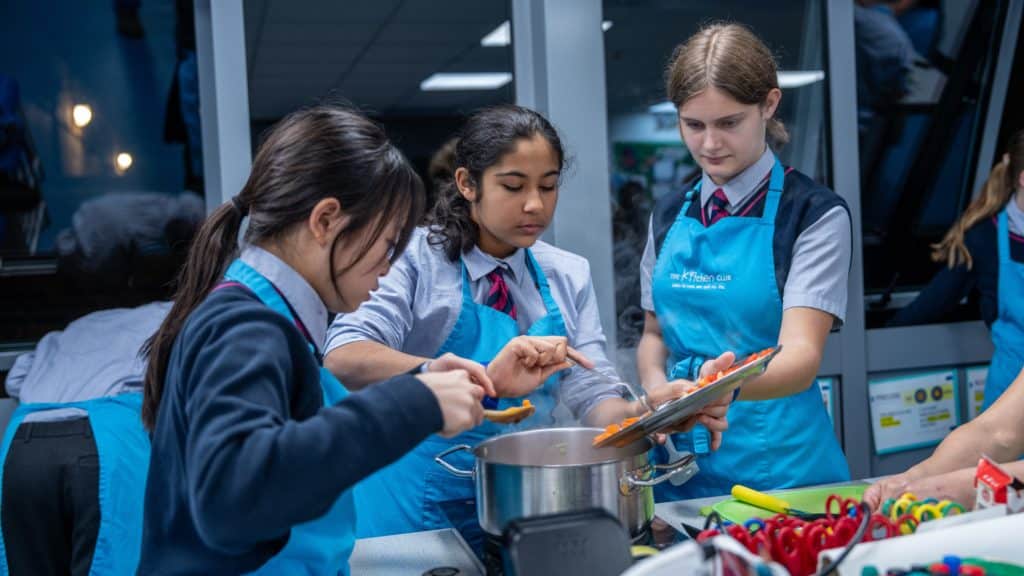Three girls from Cheltenham Prep School, wearing blue aprons and school uniforms, are cooking together, adding ingredients to a large saucepan on a kitchen worktop in a classroom setting. Cooking utensils and vegetables are visible nearby.