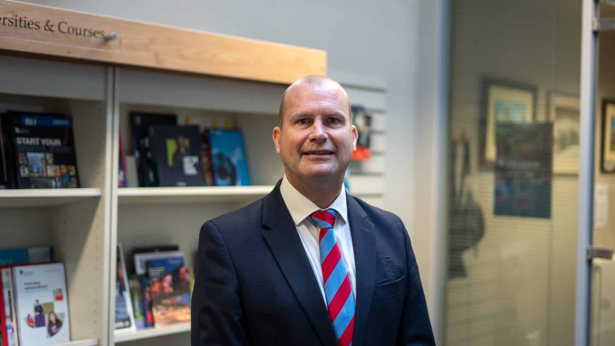 A man in a suit and striped tie stands indoors, smiling. Behind him are bookshelves filled with brochures and magazines about Cheltenham College admissions, and a glass door to the right, suggesting an office or educational environment.