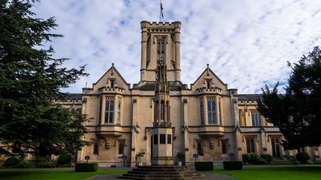 A historic stone building with a tall central clock tower, arched windows, and decorative details, Cheltenham College stands majestically amidst green trees and beneath a cloudy sky overhead.