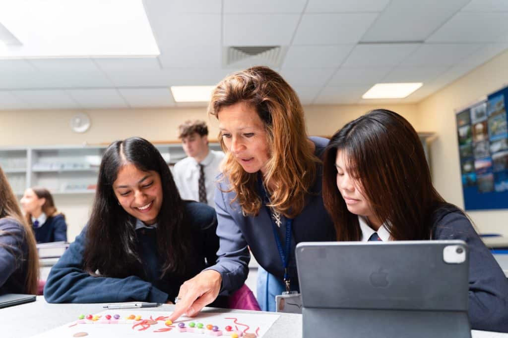 A teacher at Cheltenham College helps two pupils with a science project using colourful sweets. The pupils, wearing uniforms, smile as they work at a desk with an iPad in a bright classroom.