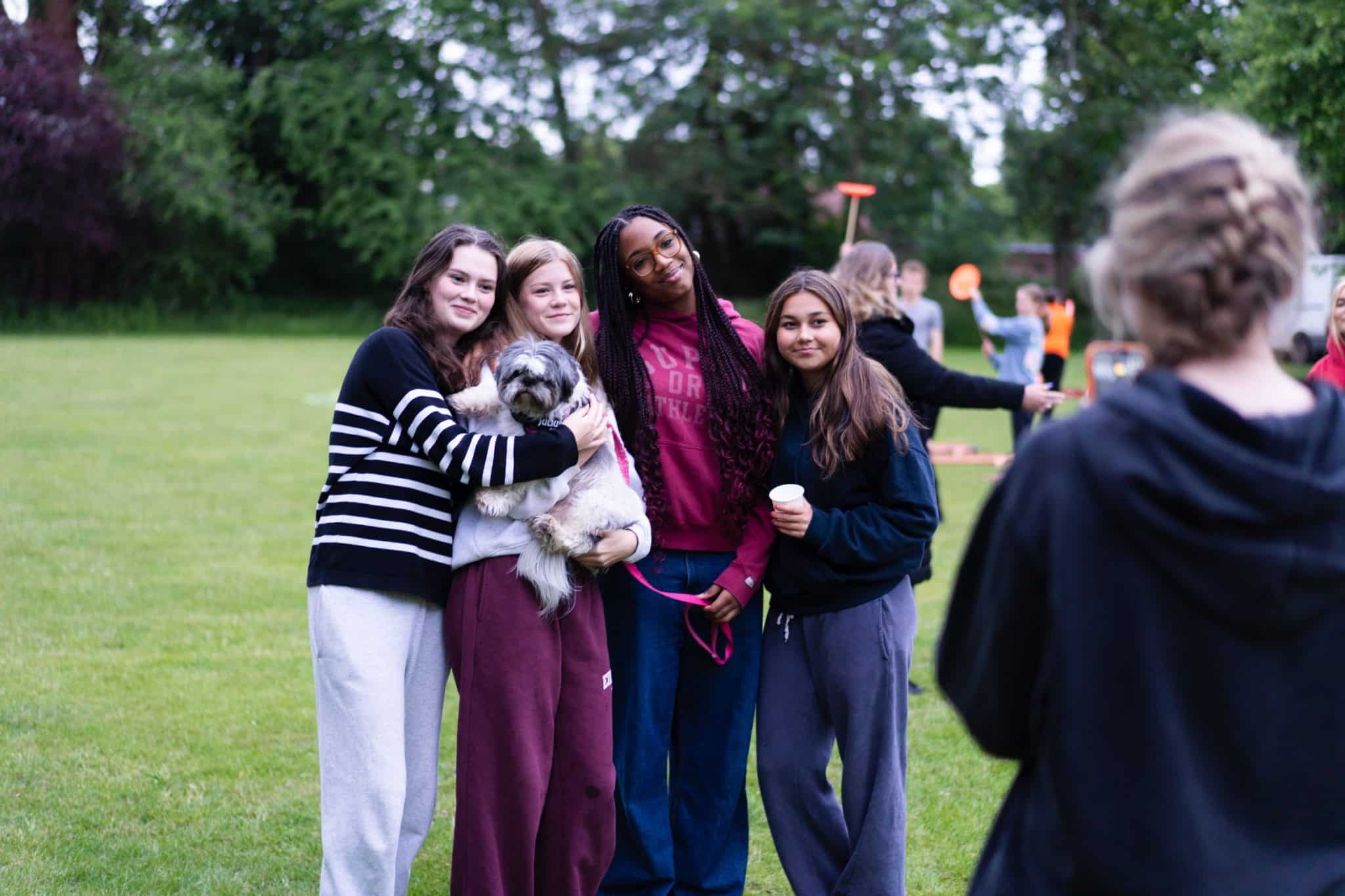 Four teenage girls stand together outdoors on grass, smiling at the camera; one holds a small fluffy dog. They wear casual clothing, and other people are visible in the blurred background. Trees surround the area.