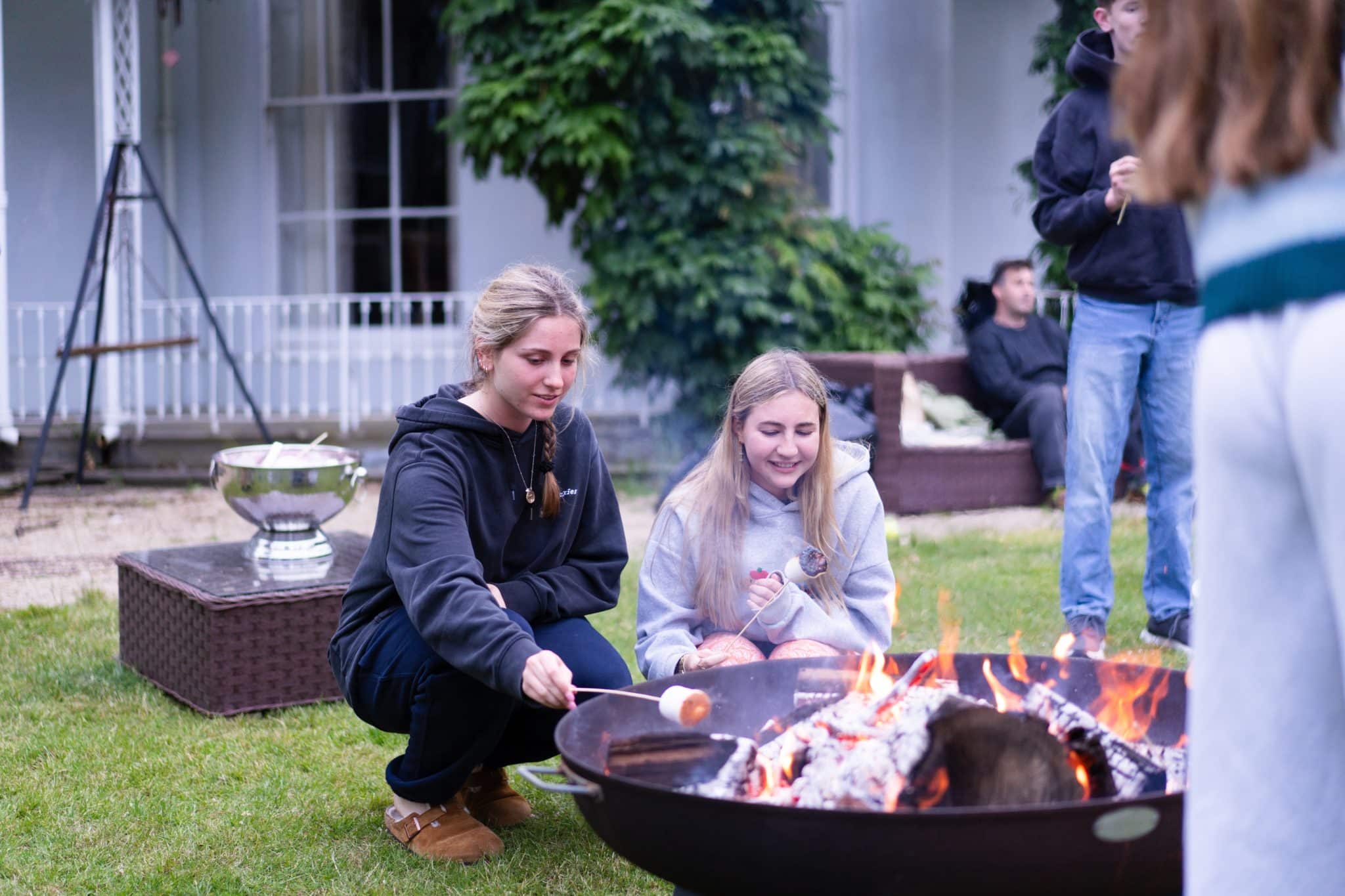 Two young women kneel by a fire pit, toasting marshmallows. Other people are gathered nearby in a grassy back garden with garden furniture and a white house in the background.