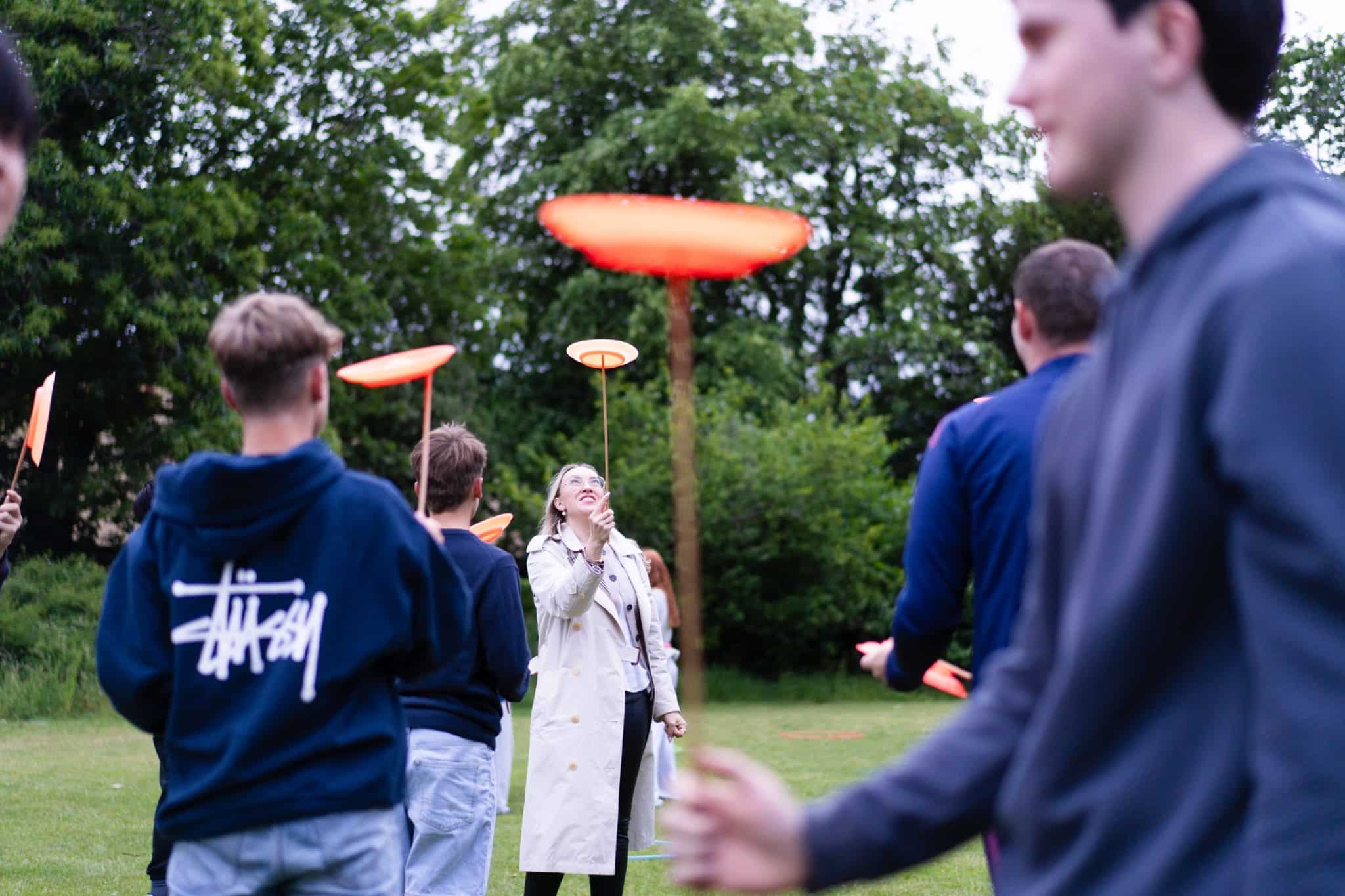 A group of people outdoors spin orange plates on sticks, with trees in the background. A woman in a beige coat smiles whilst looking up at her plate, and others focus on spinning their plates in the grassy area.