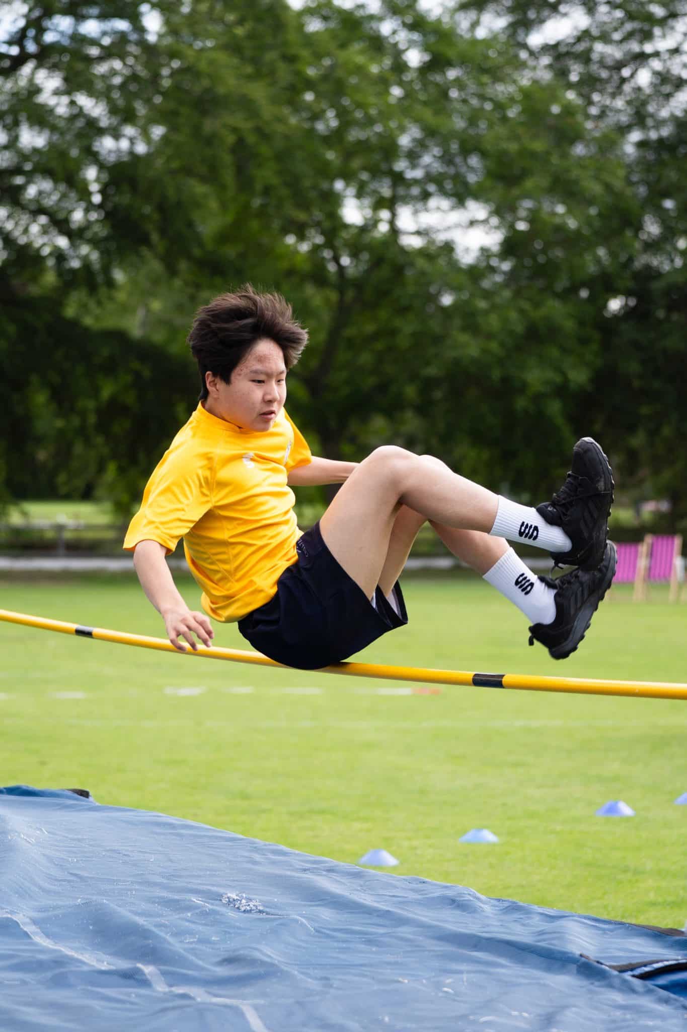 A person in a yellow shirt and black shorts is mid-air, attempting a high jump over a yellow bar on a grassy field, with trees and sports equipment visible in the background.