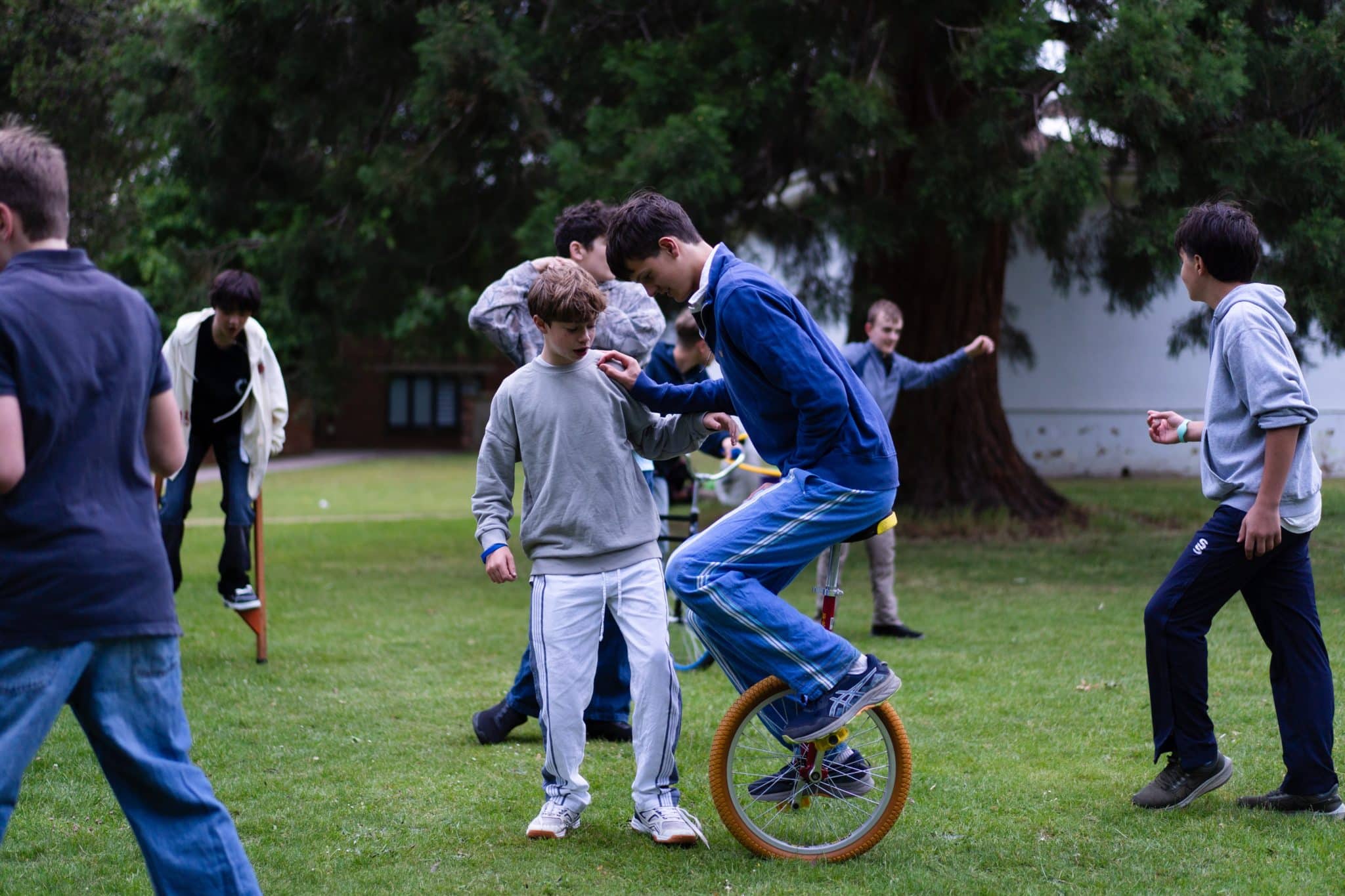A group of boys play outside on grass; one rides a unicycle whilst others walk or watch. Trees and a building are in the background, creating a lively and active atmosphere.