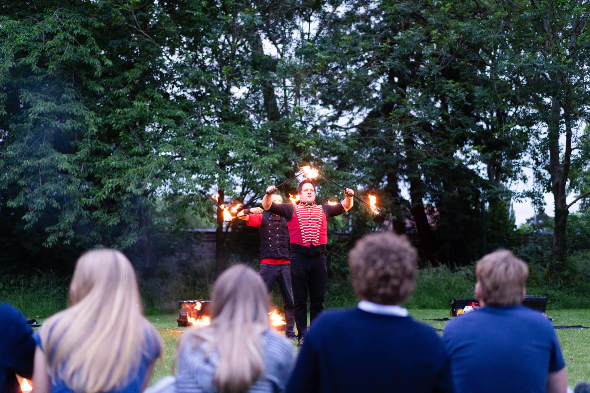 Two performers dressed in red and black juggle flaming torches outdoors, entertaining a seated audience on a grassy area surrounded by trees.