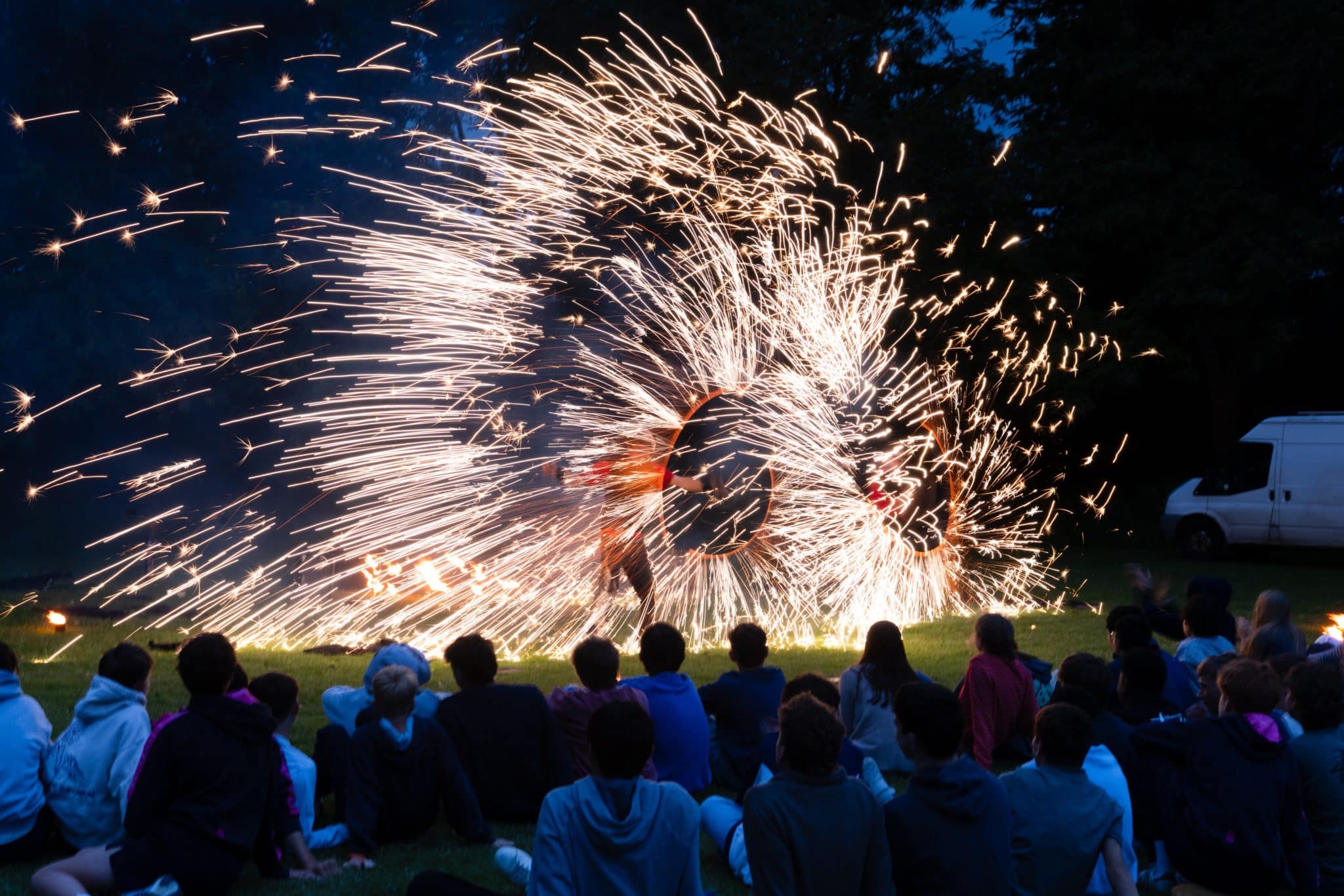 A crowd sits on grass at night watching a performer spin fire, creating bright, circular sparks in the air, with trees and a white van in the background.