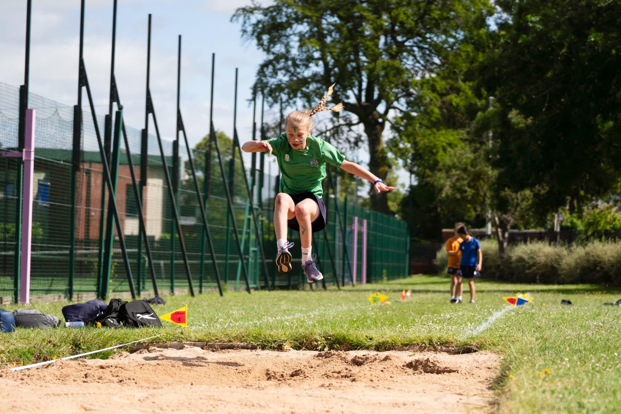 A young girl wearing a green shirt and shorts is mid-air whilst performing a long jump over a sandpit on a sports field, with sports bags and two people visible in the background.