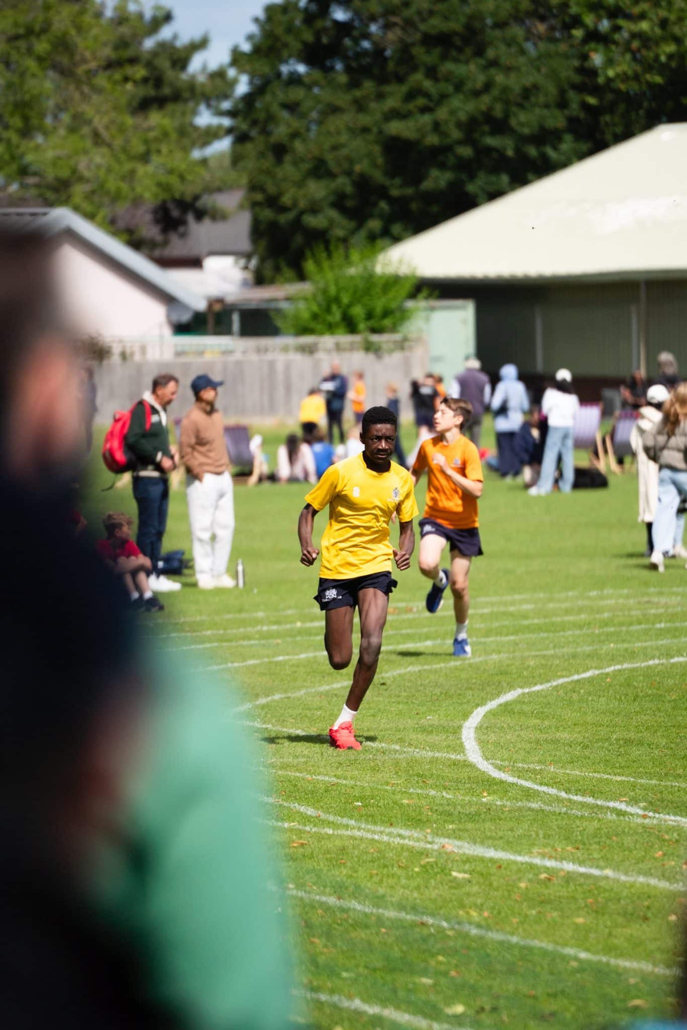 Two runners, one in a yellow shirt and one in orange, race on a grassy track. Spectators stand and watch in the background, and trees and buildings are visible. The foreground is slightly blurred by a bystander.