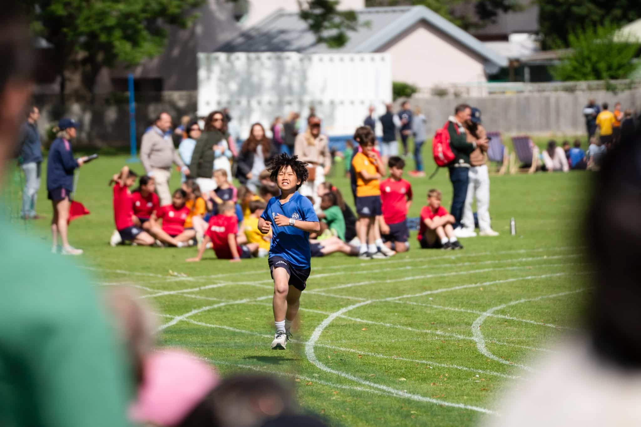 A young boy in a blue shirt runs on a grassy track during a school race, with spectators and other children watching and sitting in the background.