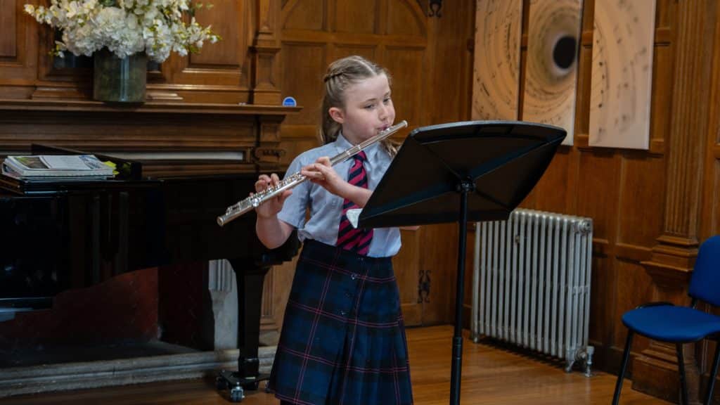A young girl in a Cheltenham College school uniform plays the flute at a music stand in a wood-panelled room with a piano, flowers, and art on the wall.