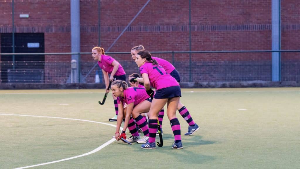 Four female hockey players in matching pink and black kits are crouched and ready at the edge of the pitch, appearing focused before play, with a brick wall in the background—a testament to the dedication seen at Cheltenham College, recognised in the Independent Schools of the Year Awards 2026.