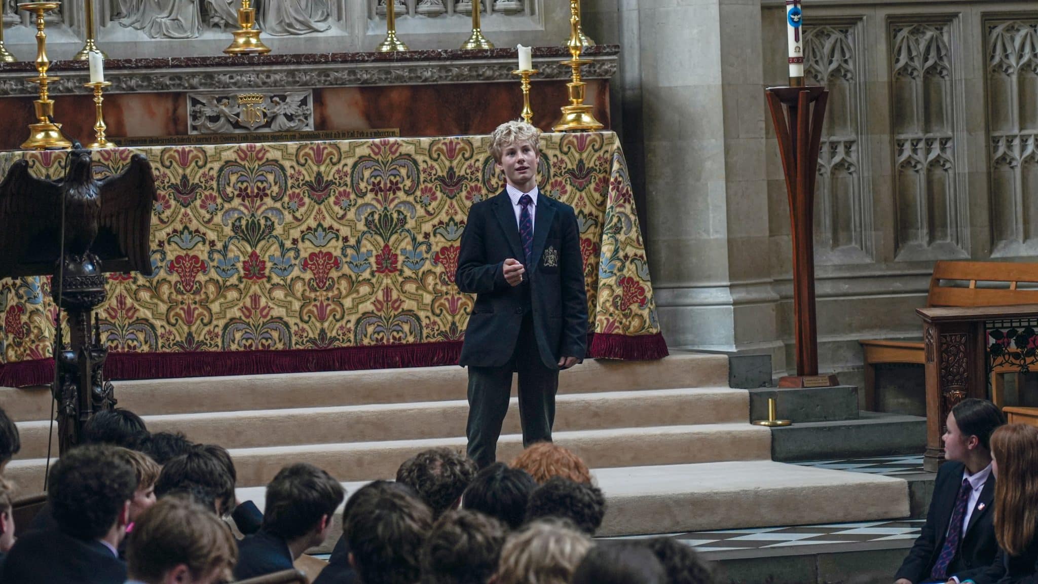 A student in a dark blazer stands speaking on the steps in front of an ornate altar in a chapel, whilst other students sit and listen. Gold candlesticks and patterned cloth decorate the altar behind him.