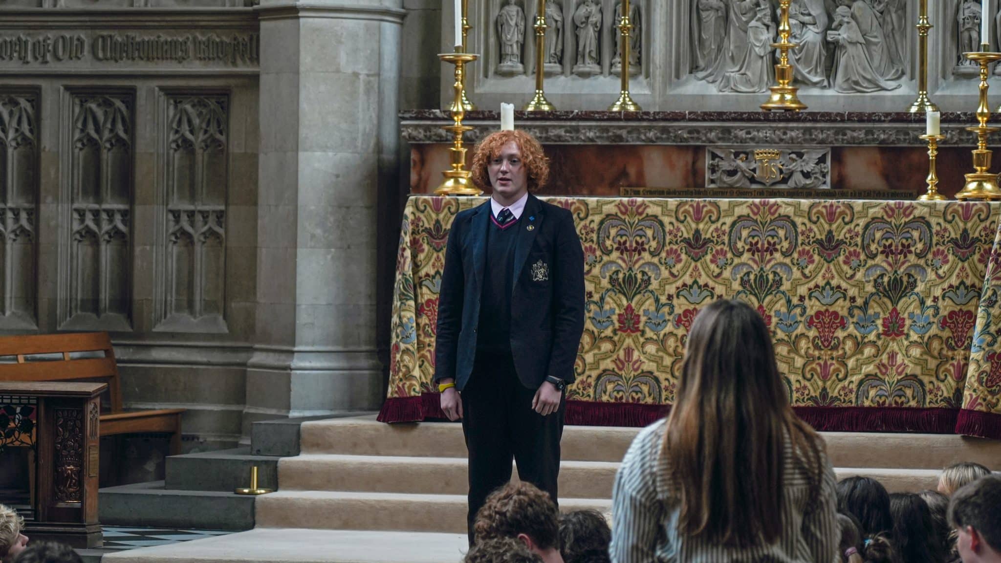 A young person with curly red hair stands on steps in front of an ornate altar in a church, wearing a dark blazer and tie, whilst an audience looks on. A woman with long hair is in the foreground facing him.