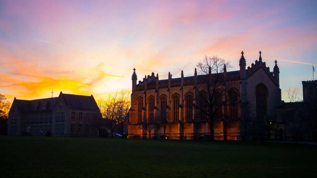 A large, gothic-style building at Cheltenham College is lit by a colourful sunset sky. The silhouette of leafless trees stands in front, with another old building on the left and green lawn in the foreground.