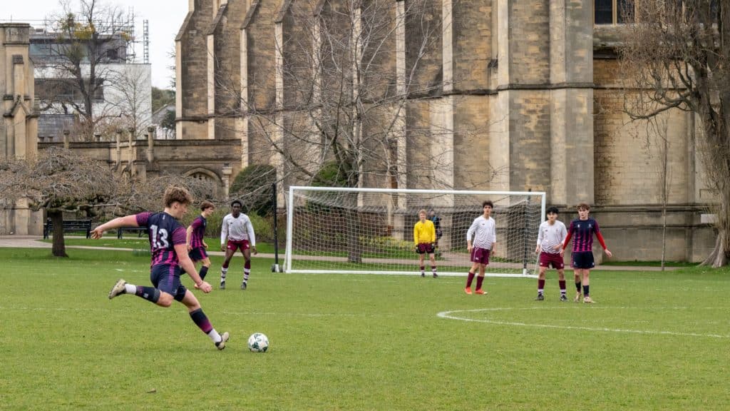 A footballer in a navy and pink kit prepares to kick the ball while others watch near a goal on a grassy pitch beside a large stone building at Cheltenham College, a proud participant in the Independent Schools of the Year Awards 2026.
