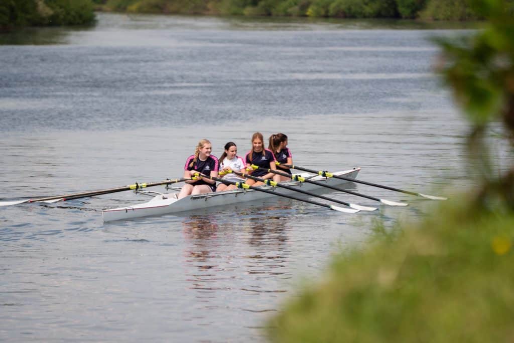 Four girls in a rowing boat paddle together on a calm river, surrounded by greenery. Wearing matching sports kit, they appear focused as they work as a team—demonstrating the discipline and collaboration often fostered through Oxbridge Preparation.
