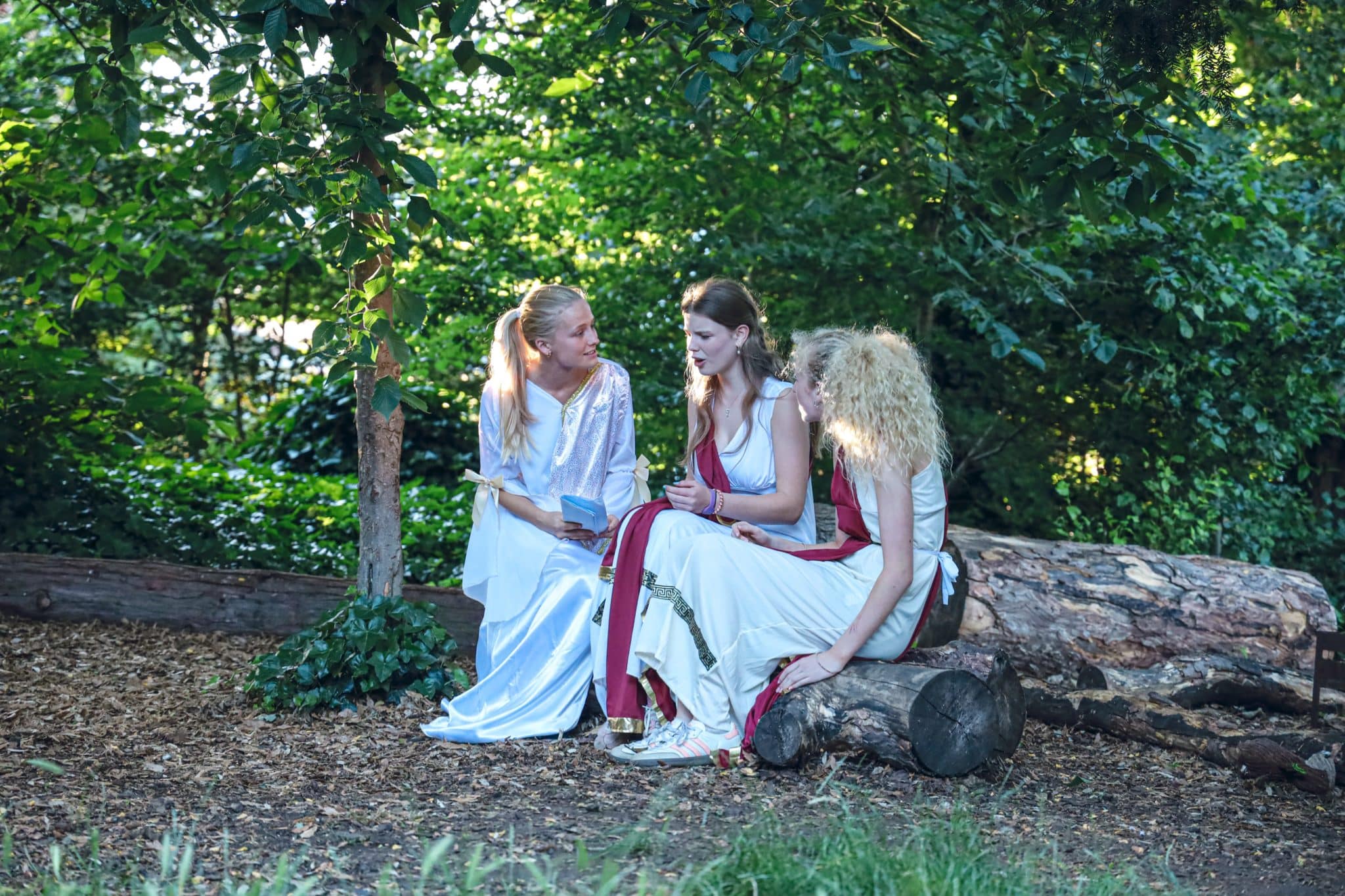 Three women in long, flowing white dresses sit on logs in a wooded area, talking and smiling together in the shade of trees. Sunlight filters through the leaves, creating a peaceful, natural setting.