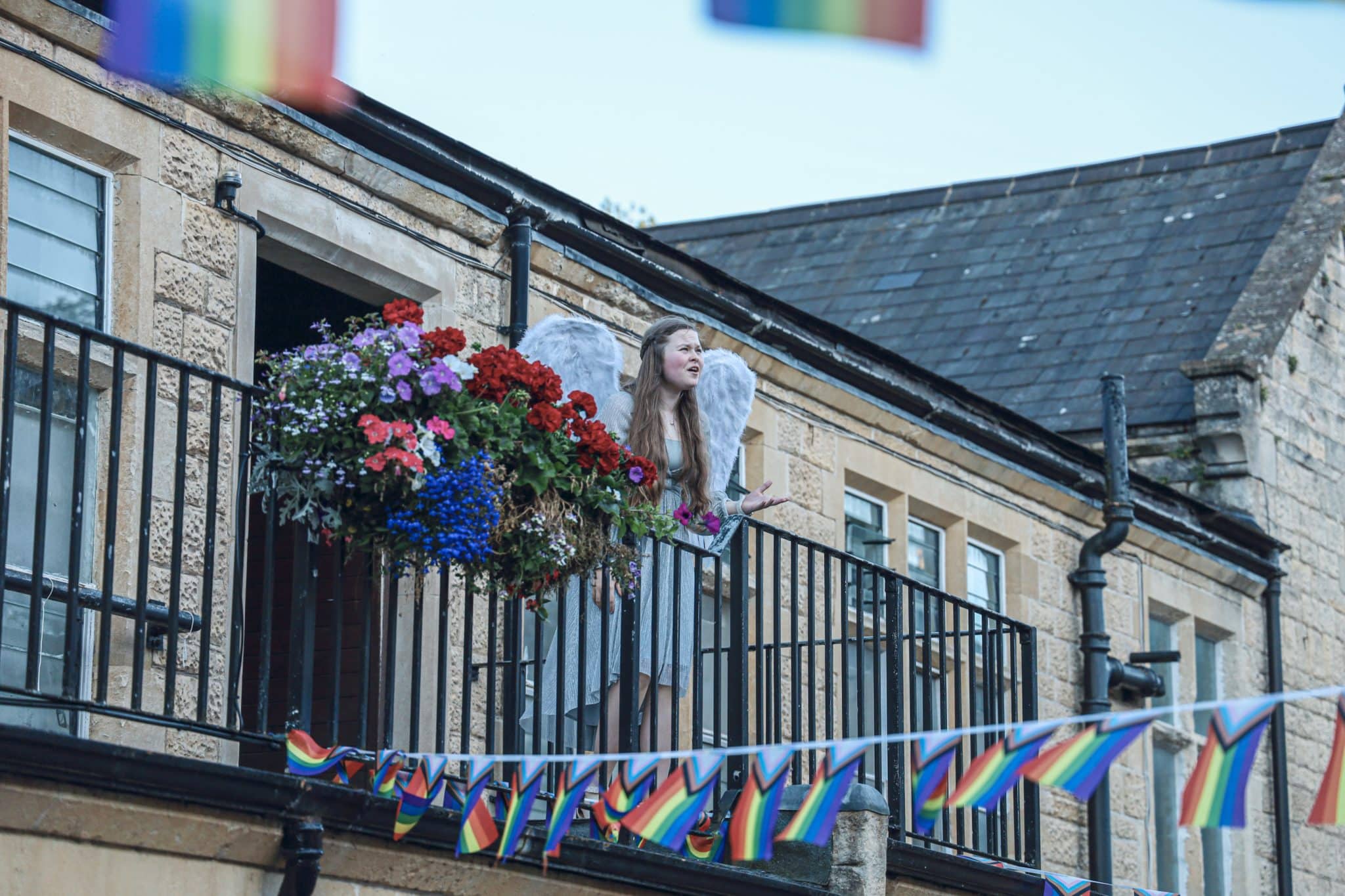 A person dressed as an angel stands on a balcony decorated with colourful flowers. Pride flags hang below and in the foreground, and the building has tan stone walls and black railings.