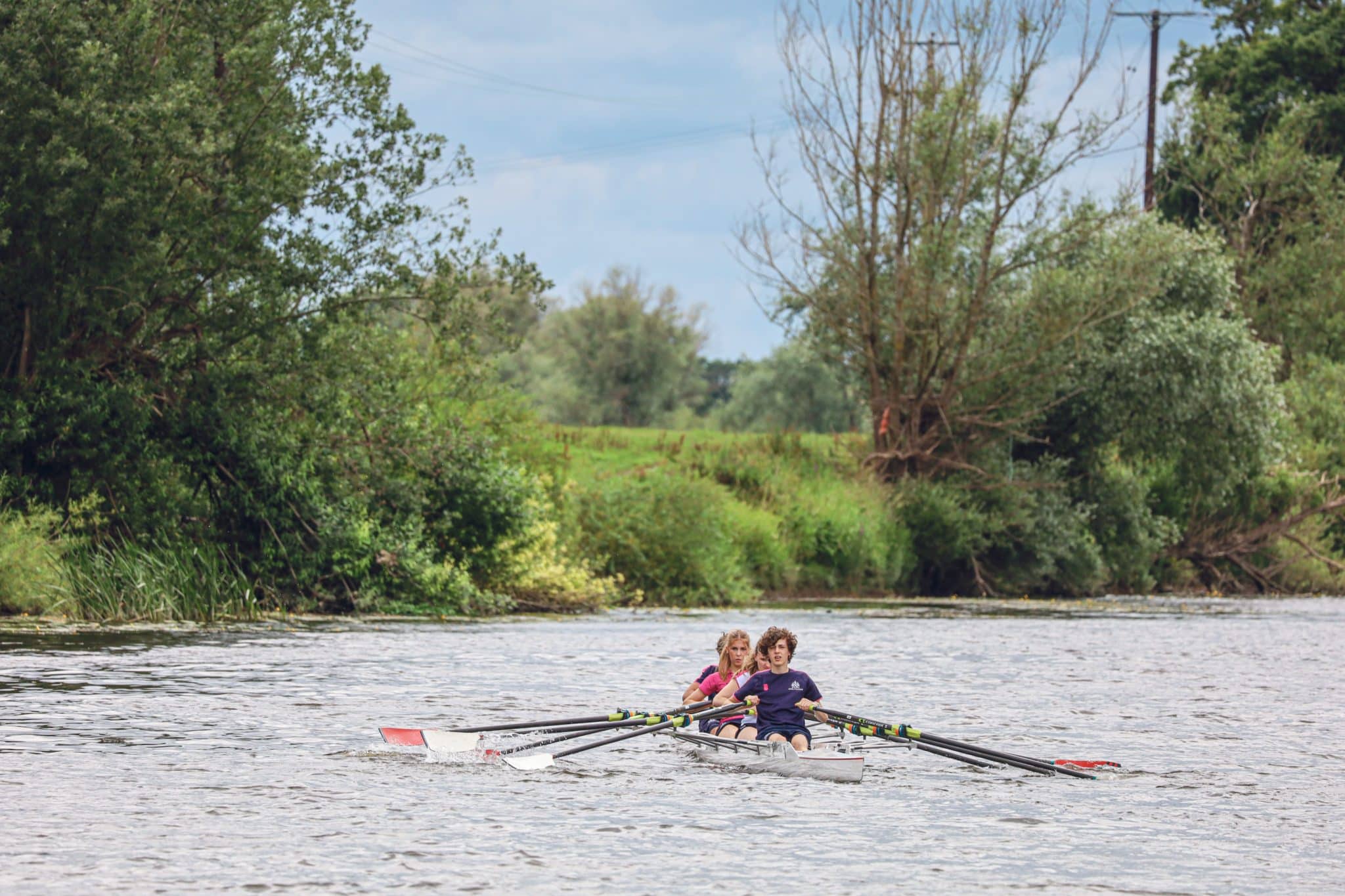 Three people are rowing together in a boat on a river, surrounded by lush green trees and vegetation on the riverbank under a cloudy sky.