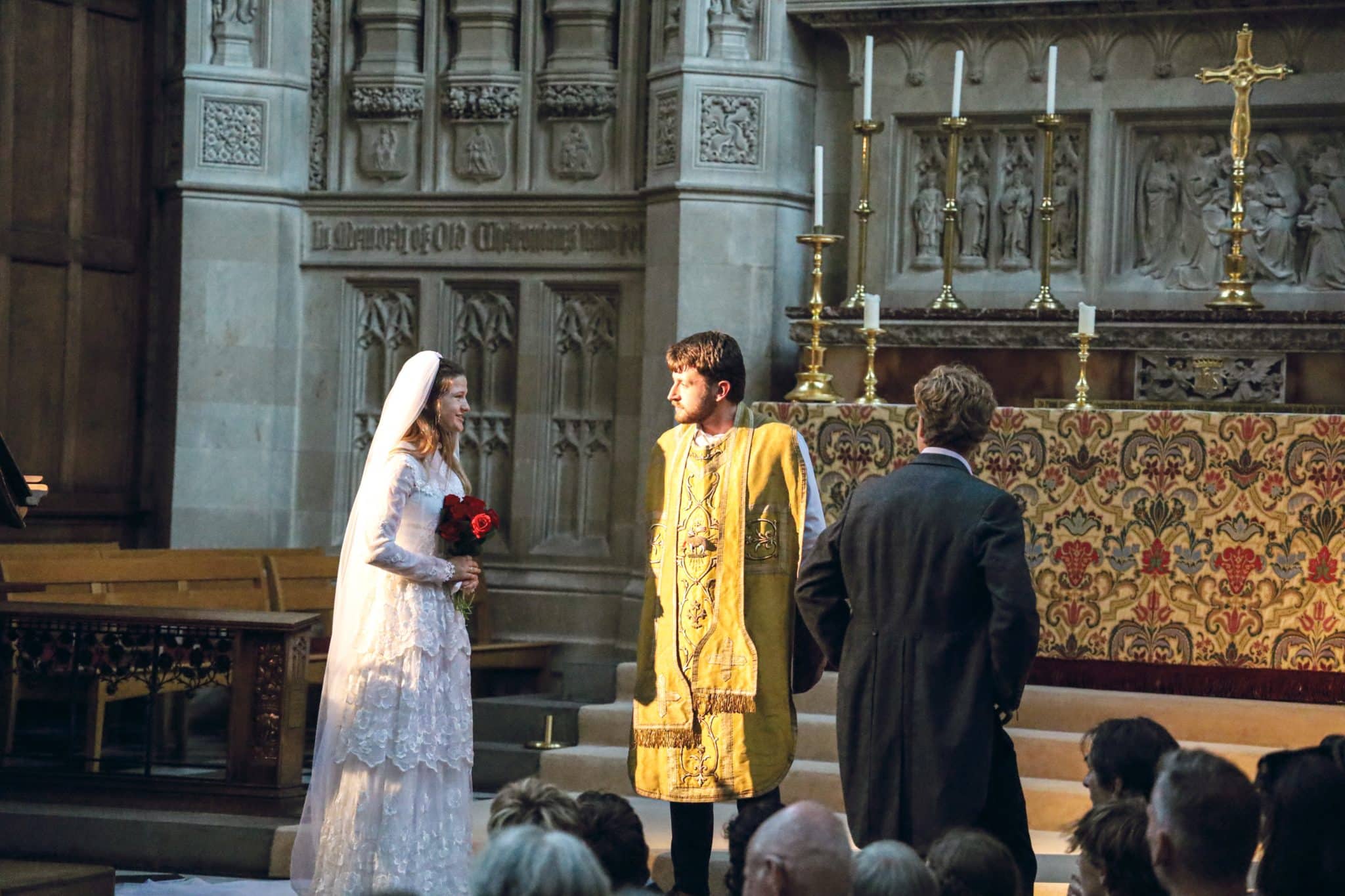 A bride in a white dress and veil holding red flowers stands next to a groom in ornate gold robes before an altar in a church, with another man in a suit standing nearby. An audience sits facing them.