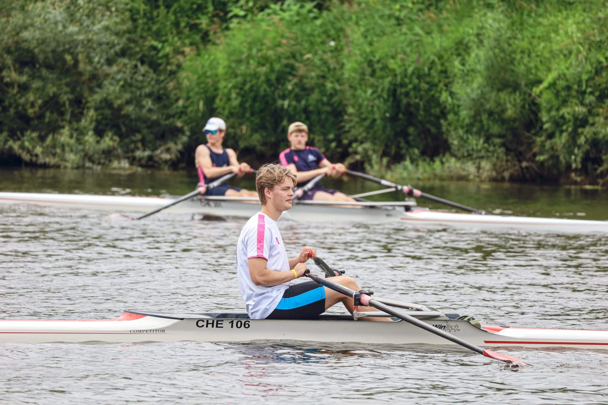 A man in a single scull rows on a river, with two other rowers in a double scull behind him. All are wearing sports kit, and green trees line the riverbank in the background.
