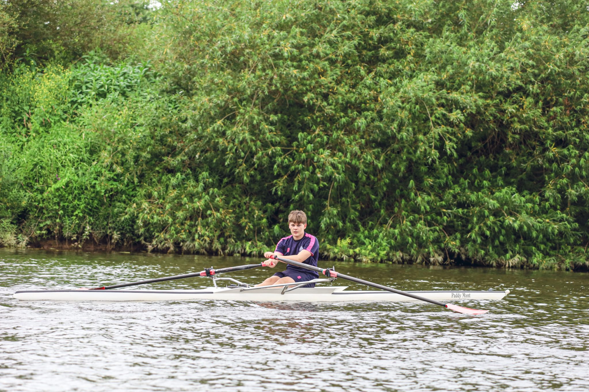 A person is rowing a single scull boat on a calm river, with green, leafy bushes in the background and cloudy skies overhead.