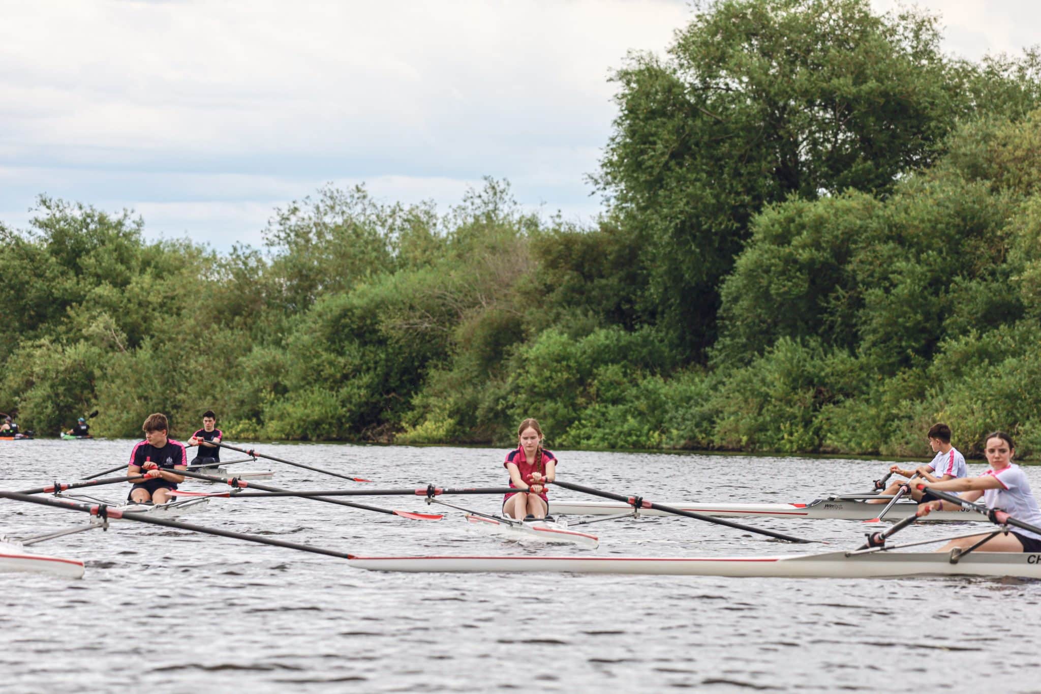 A group of young people rowing in single sculls on a river, surrounded by green trees and bushes under a cloudy sky.