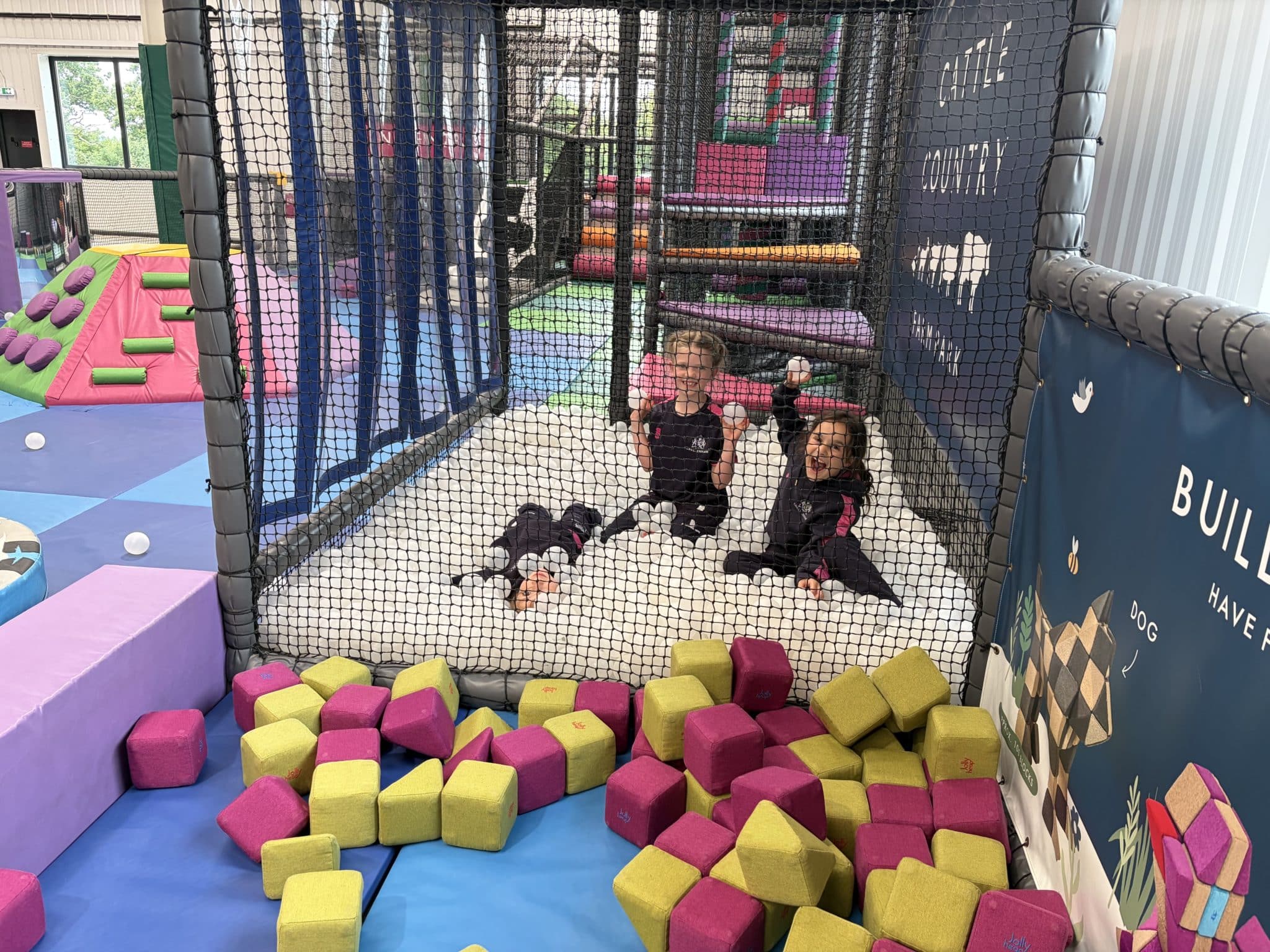 Three children sit and smile inside a netted ball pit filled with white balls at an indoor play centre. Colourful foam blocks are scattered outside the net, and play structures are visible in the background.