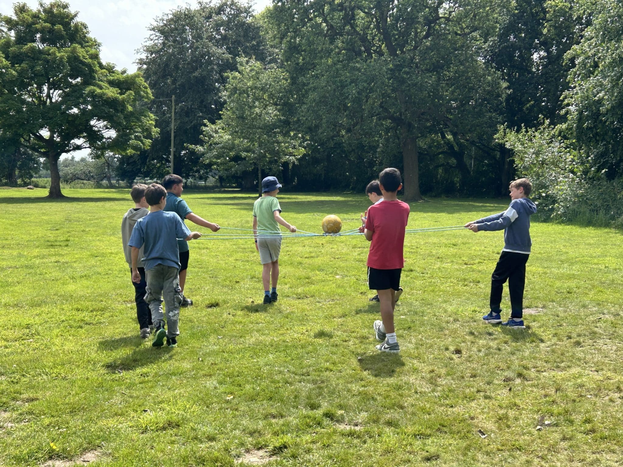 Seven children stand in a grassy field, holding a rope in a circle with a yellow ball balanced in the middle. They appear to be working together outdoors under trees on a sunny day.