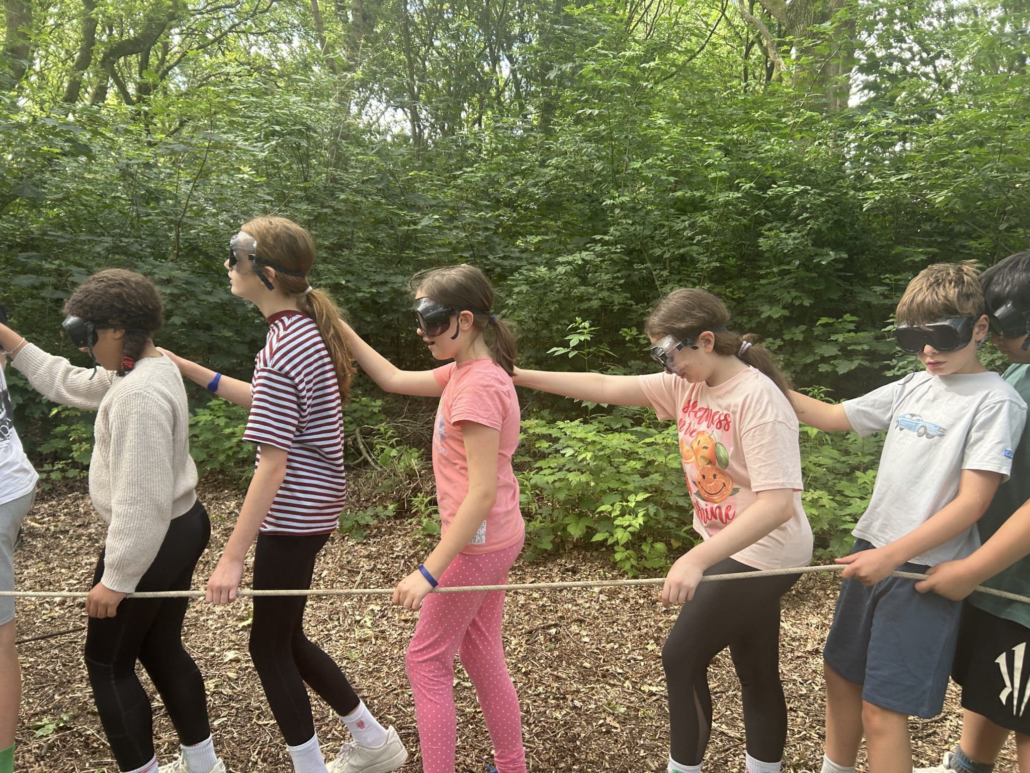 A group of children wearing blindfolds walk in a line outdoors, each holding onto the shoulder of the person in front of them and following a rope. They are surrounded by green trees and foliage.