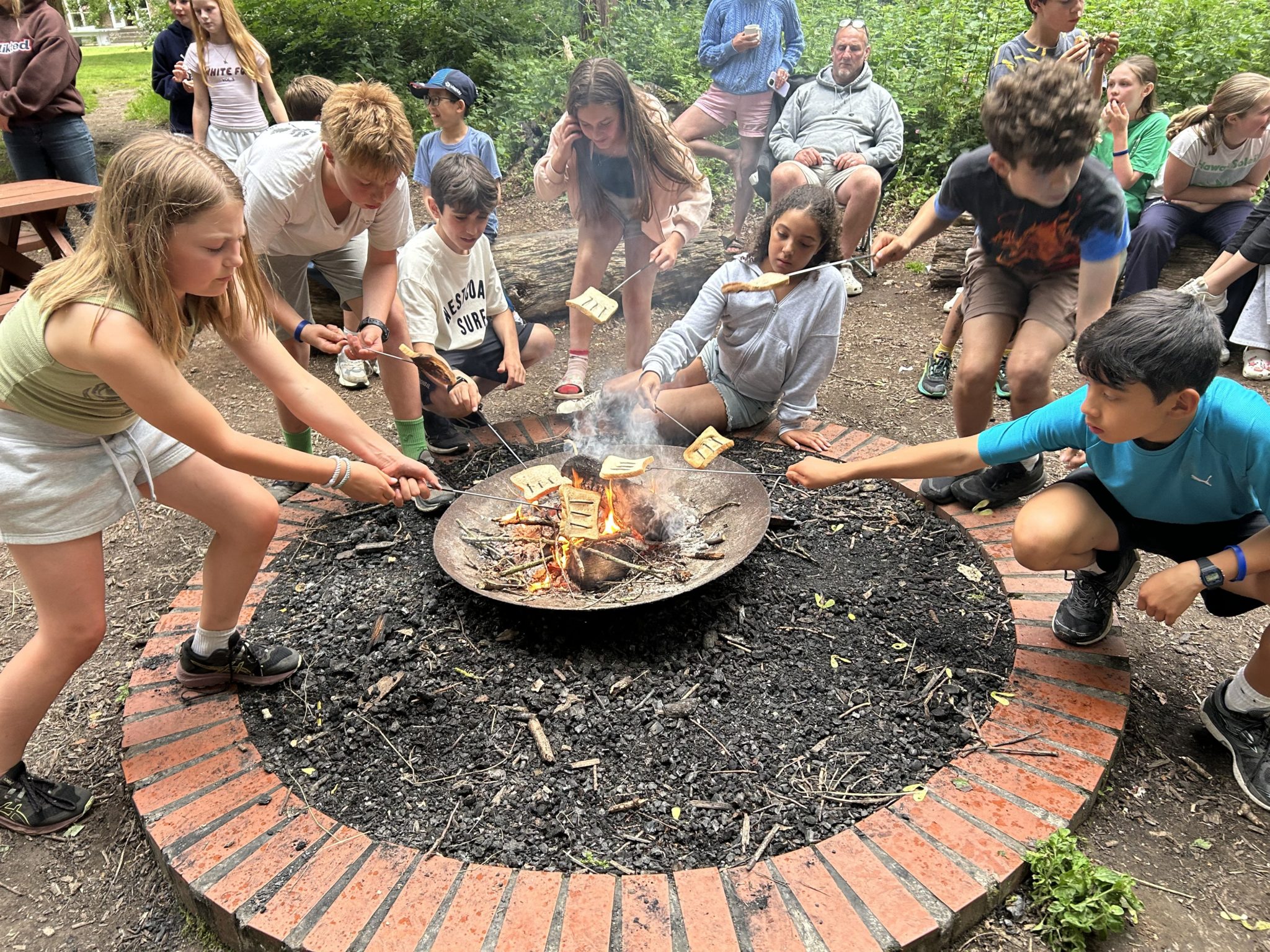 A group of children crouch and lean over a fire pit, toasting bread and marshmallows on sticks, while adults and other kids sit and watch in the background at an outdoor gathering.