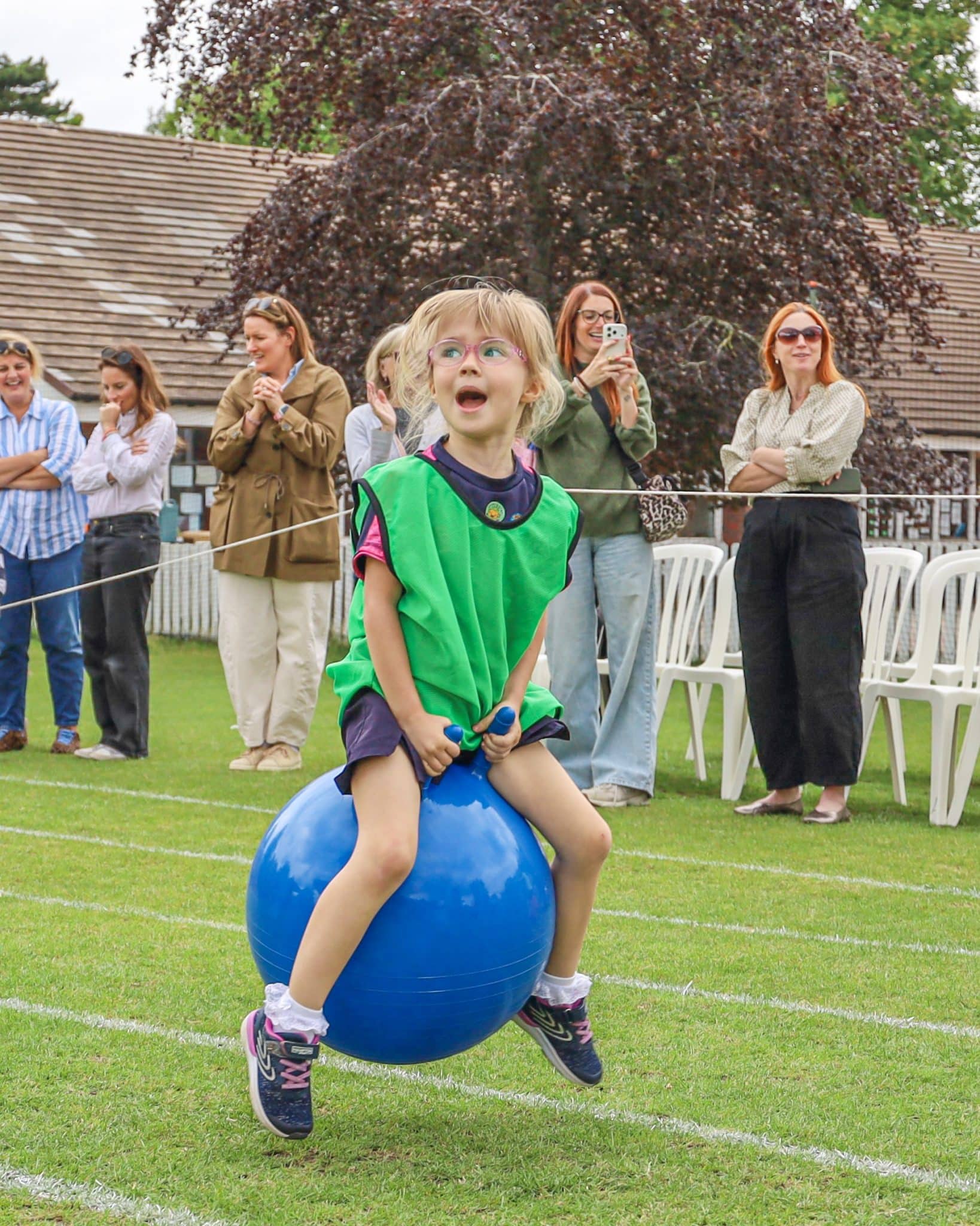 A young child in a green sports bib bounces on a blue space hopper during an outdoor event, with adults standing and watching in the background on a grassy field.