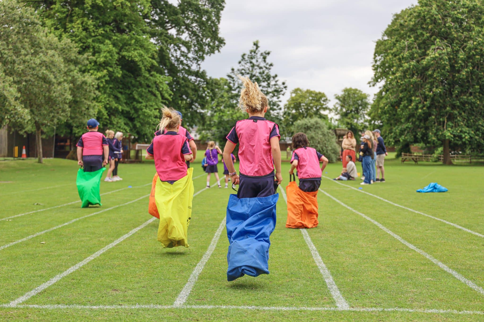 Children wearing colourful sacks participate in a sack race on a grassy field, with trees and spectators in the background on a cloudy day.