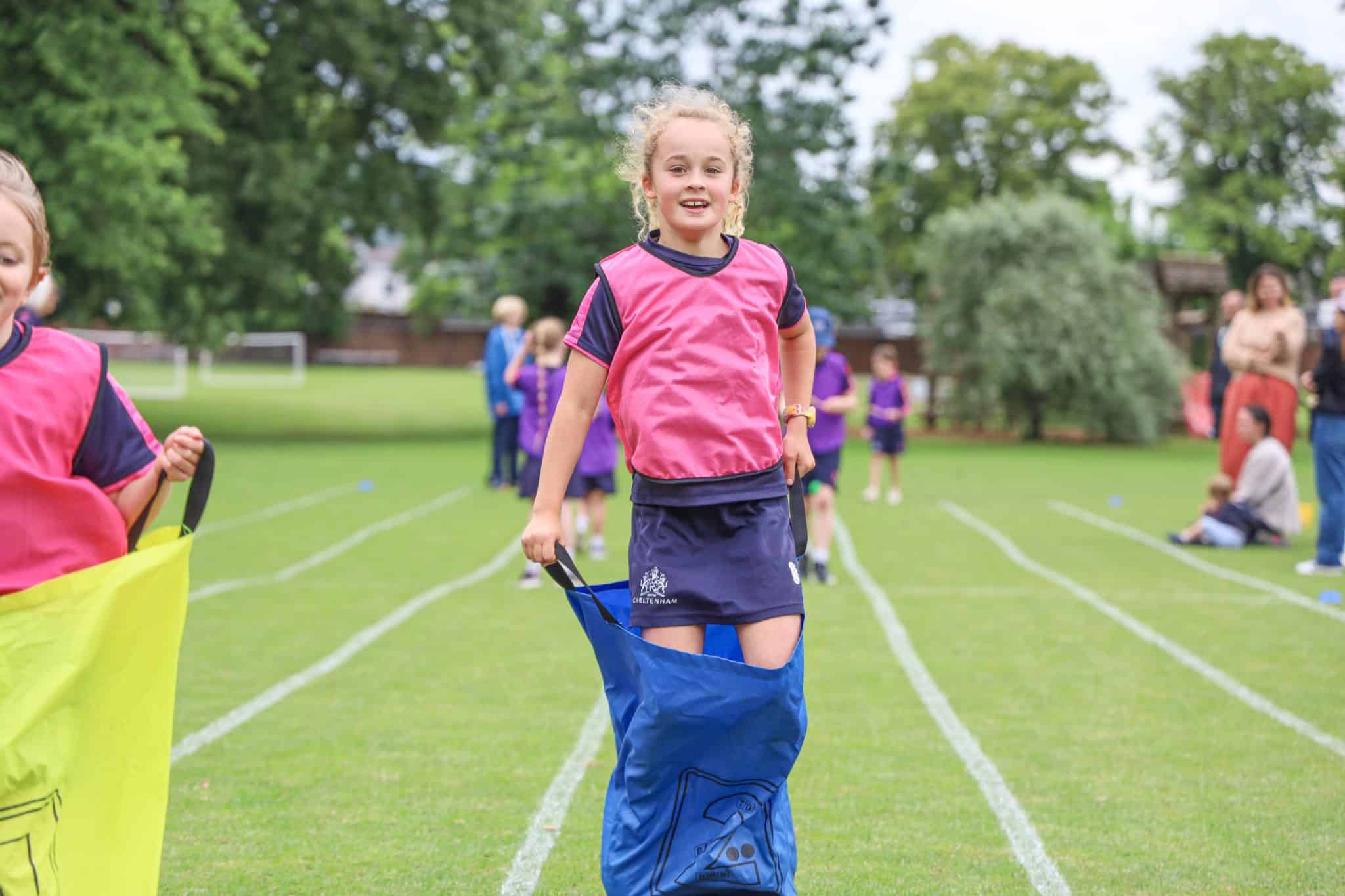 A young girl wearing a pink sports bib and blue sack takes part in a sack race on a grassy field, with other children and adults in the background.