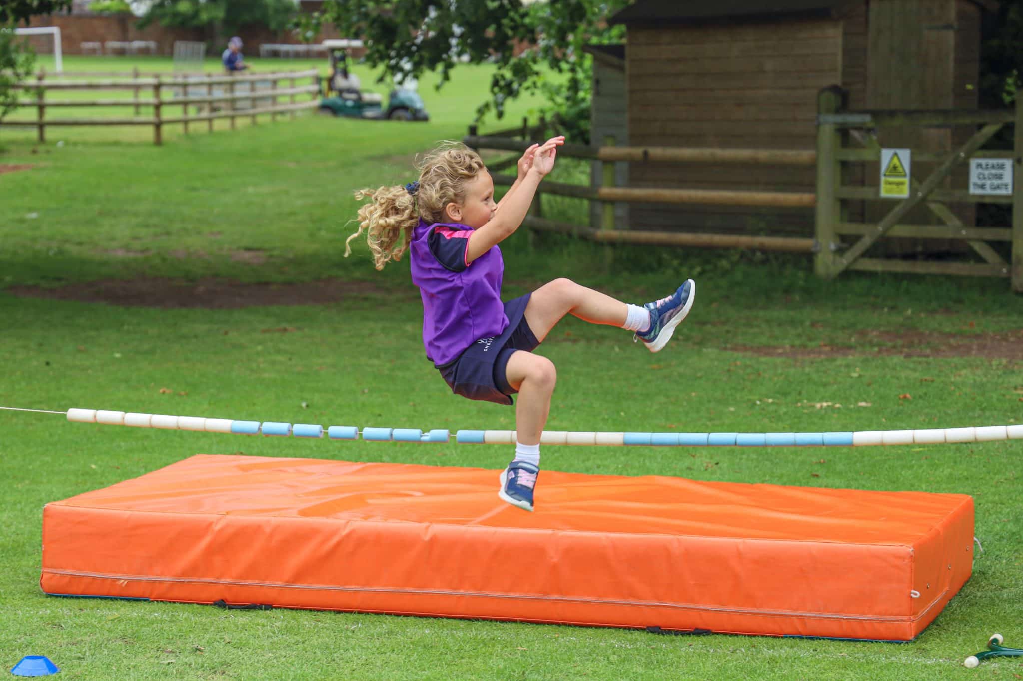 A young child with curly hair and a purple shirt is mid-jump over a bar during a high jump activity, landing onto an orange mat on a grassy field, with a wooden fence and building in the background.