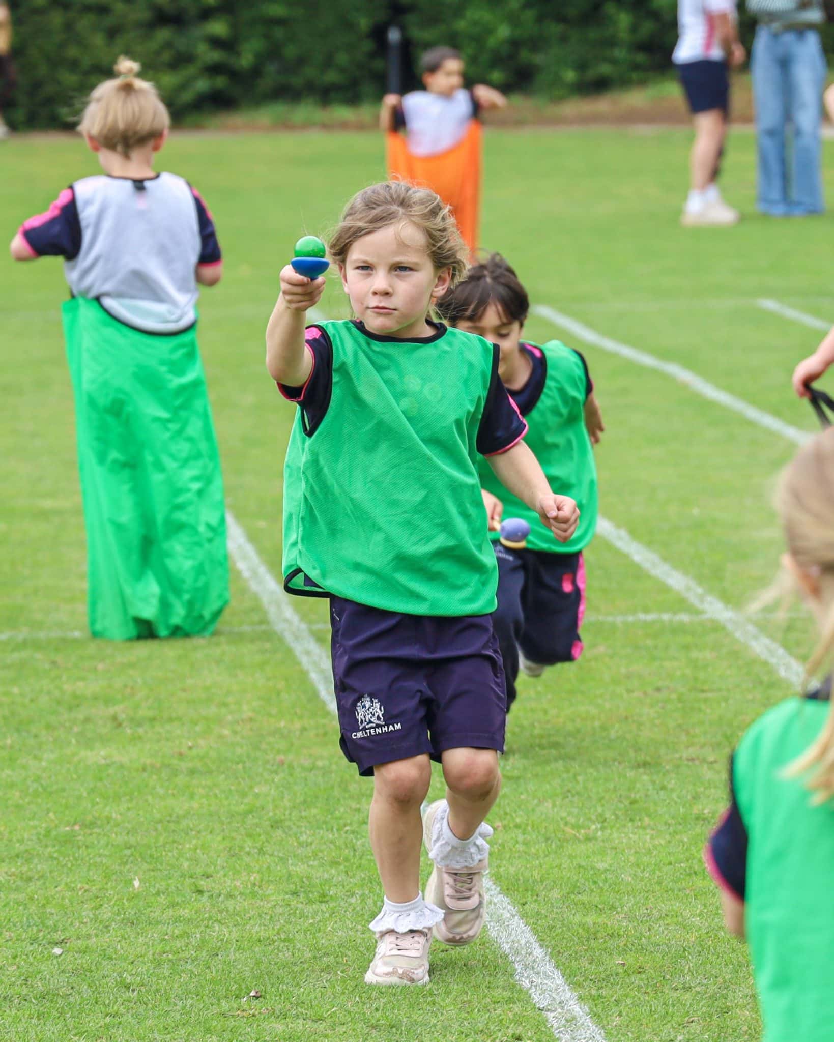 A young girl wearing a green pinnie and dark shorts runs on a grassy field holding a spoon with a ball, taking part in an outdoor sports day event with other children in the background.
