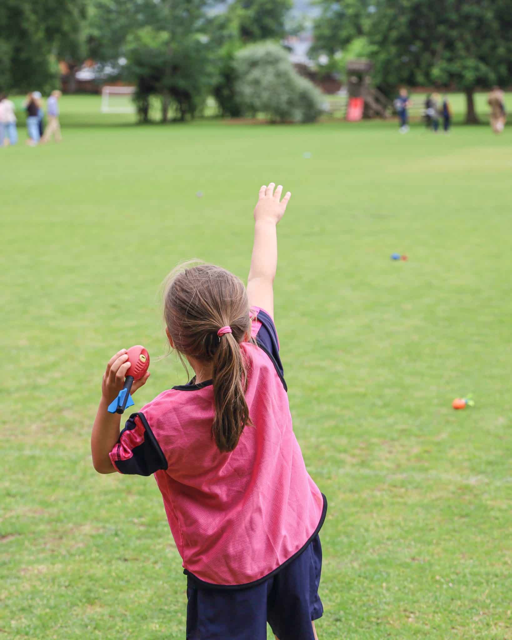 A young child in a pink sports bib stands on a grassy field, preparing to throw a small red ball with one arm extended forwards. Other people and scattered objects are visible in the background.