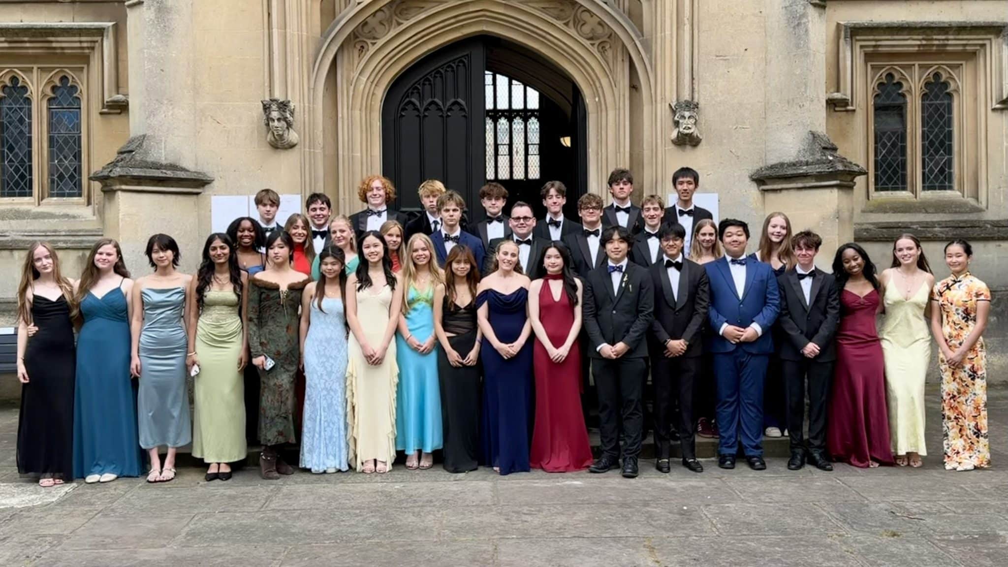 A group of young adults dressed in formal attire pose for a photo in front of a large, ornate stone building with arched doors and gothic windows.