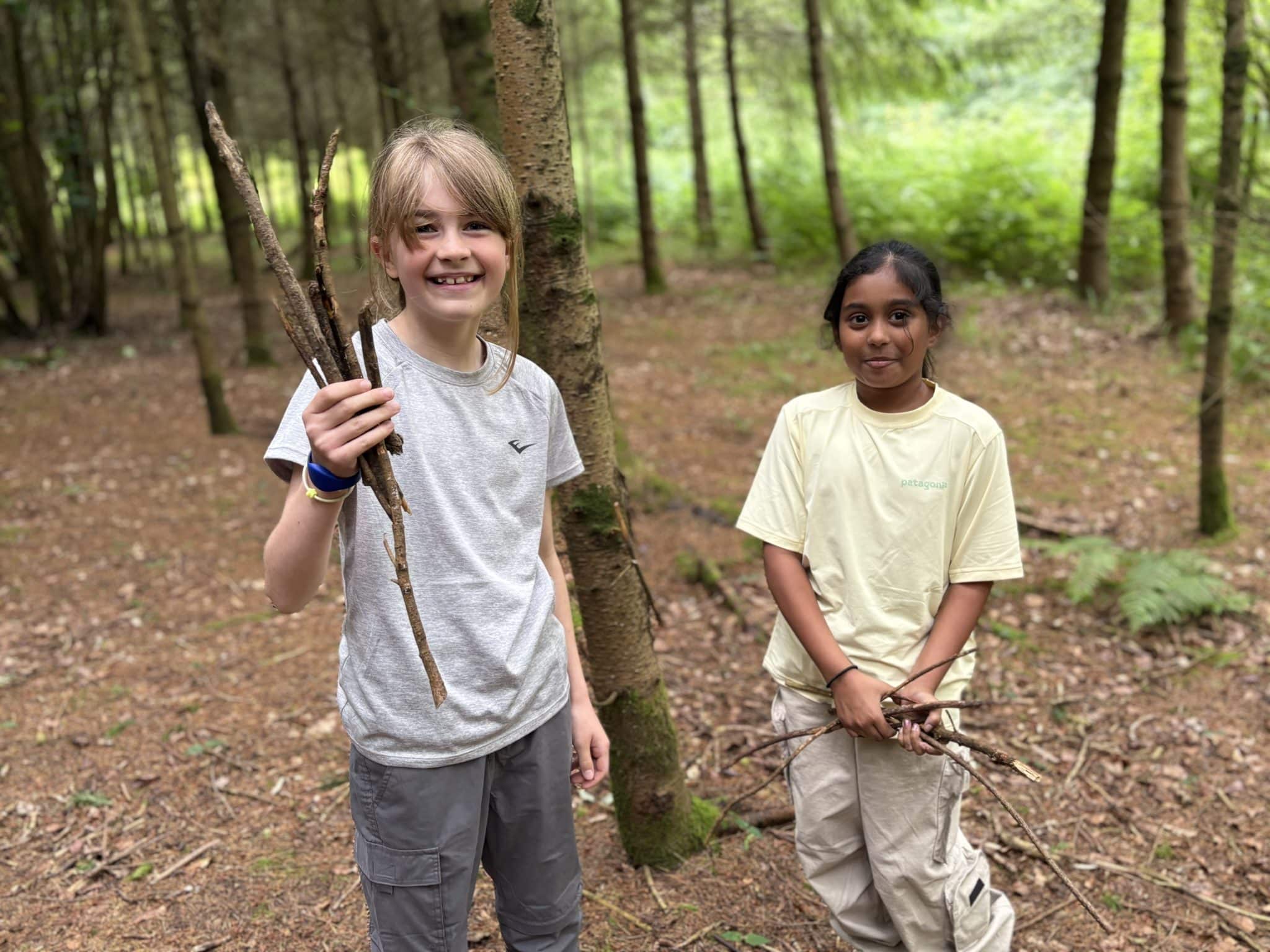 Two children enjoy their Year 4 Bushcraft Adventure at Cheltenham Prep, standing in a forest and smiling. One child holds several sticks in their right hand, whilst the other holds sticks in both hands. Green trees and foliage fill the background.