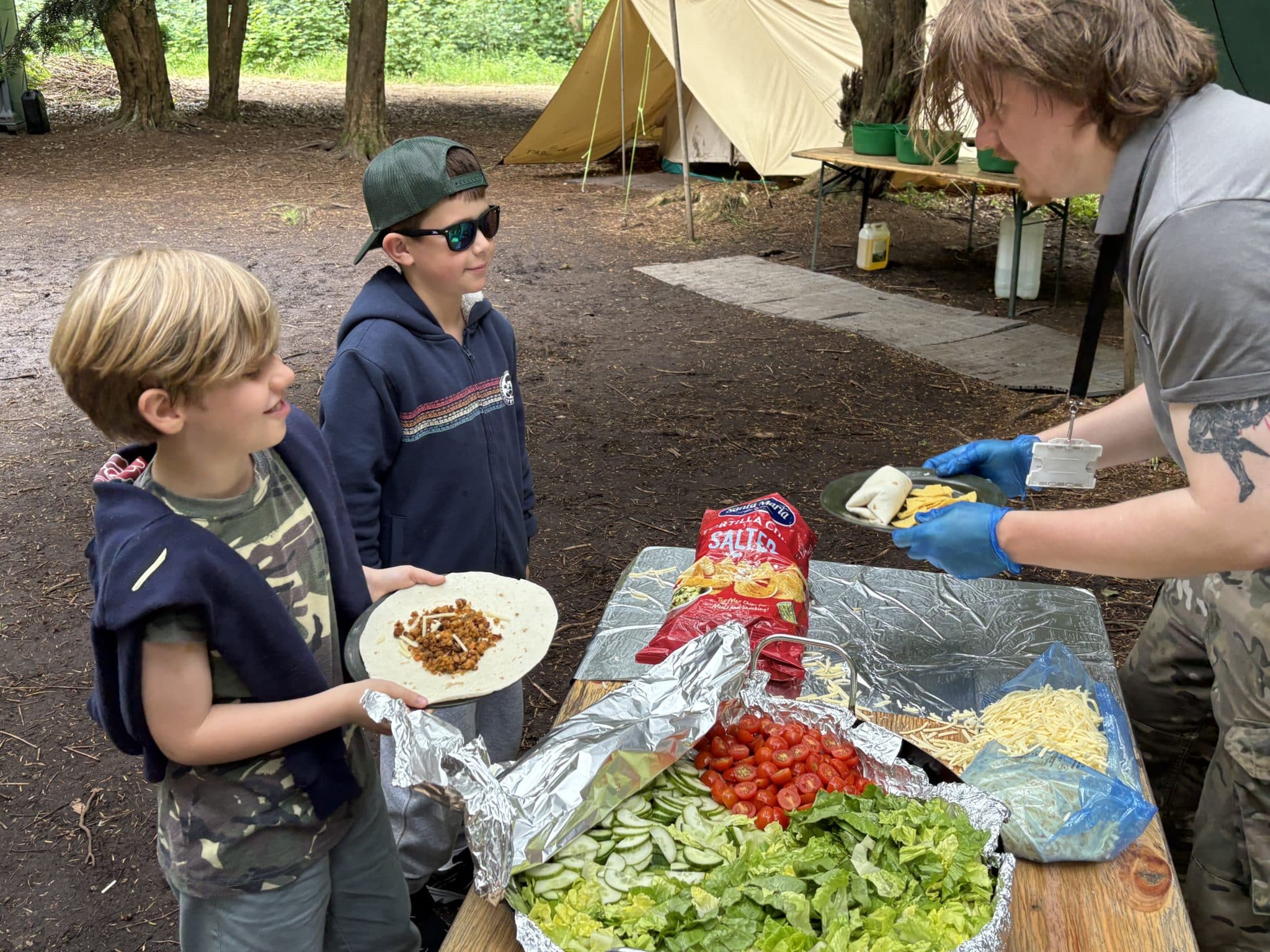 Two young boys stand outdoors by a table with salad, cheese, and crisps, holding tortillas as an adult serves food during the Year 4 Bushcraft Adventure at Cheltenham Prep. A tent and trees are visible in the background.