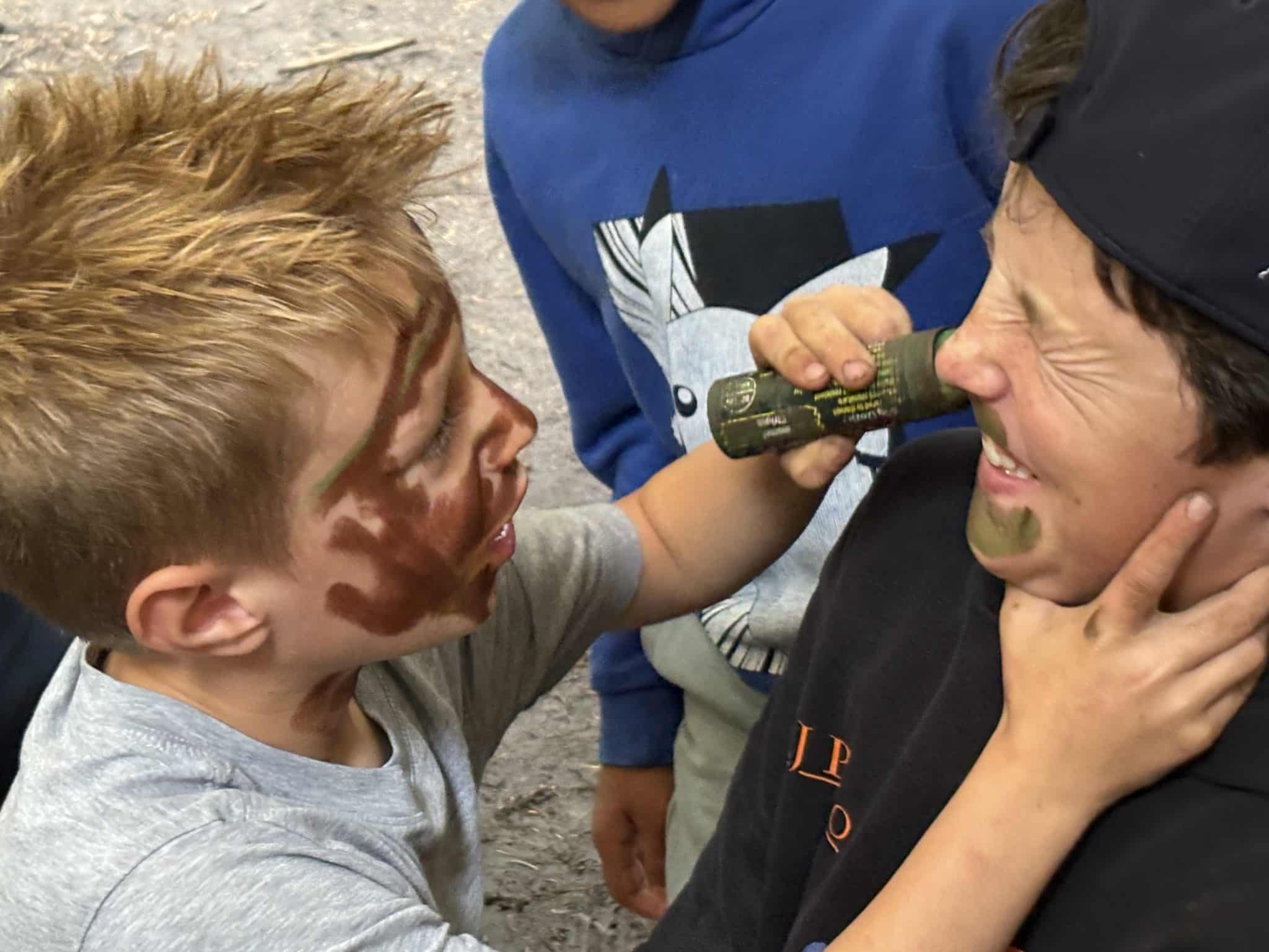 A child applies green and brown face paint to another child's face, who is squinting and smiling. Both children appear playful and happy, surrounded by others outdoors.