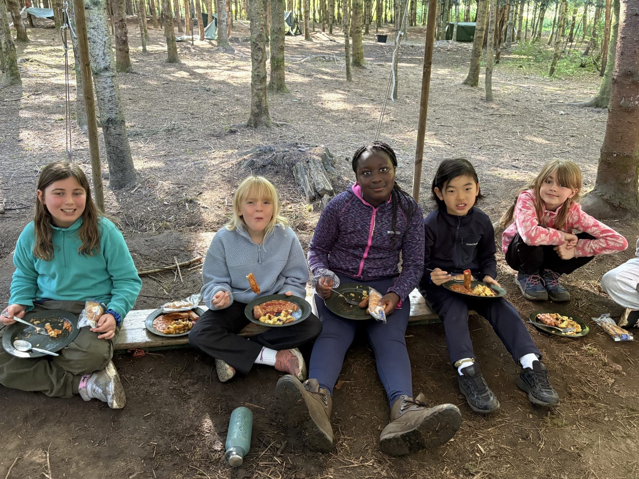 Five children sit on a wooden bench eating food from plates in a forested area. They are dressed in casual outdoor clothing and appear to be enjoying a meal together at a campsite.